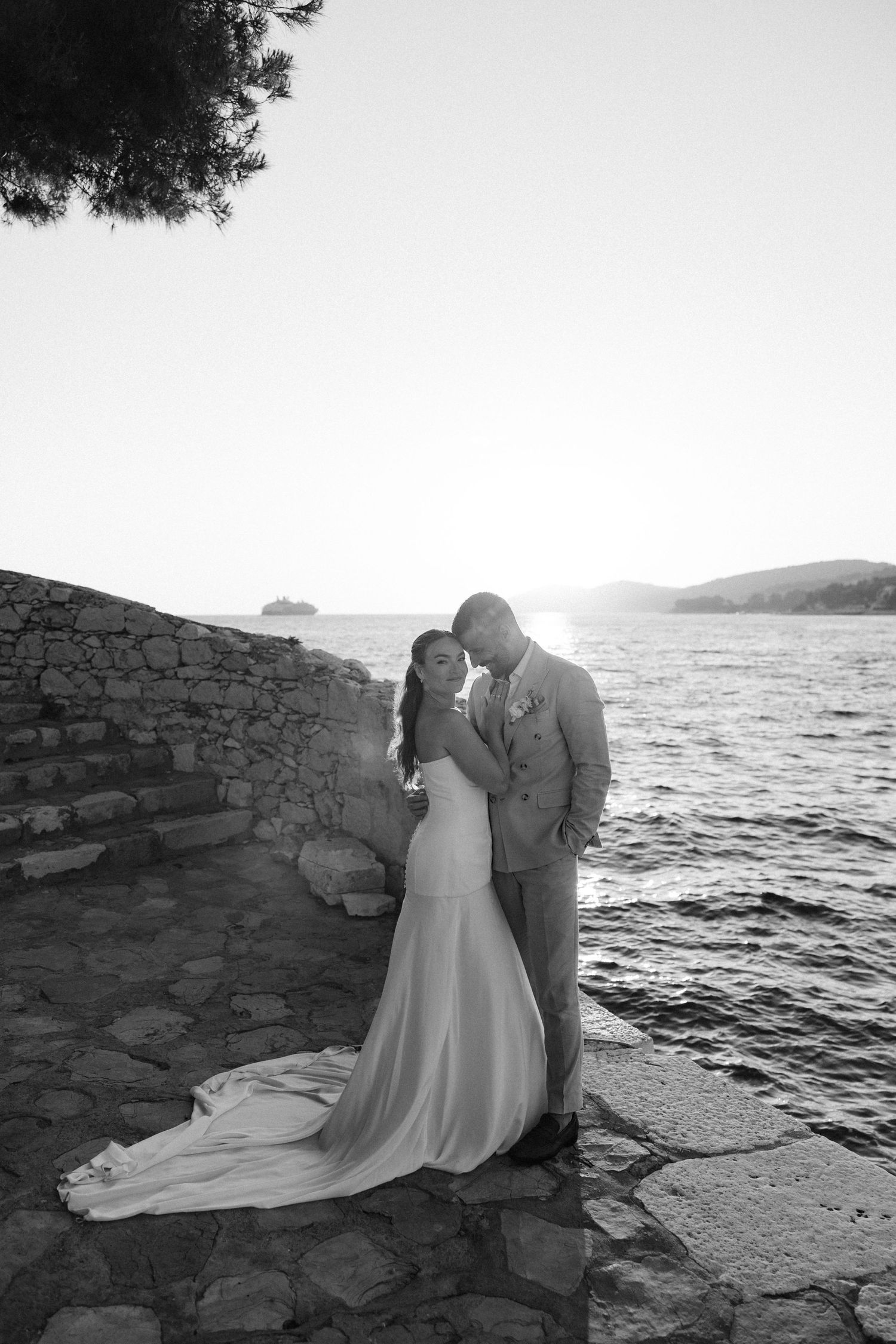Black and white photo of bride and groom embracing by the sea at sunset on stone steps.