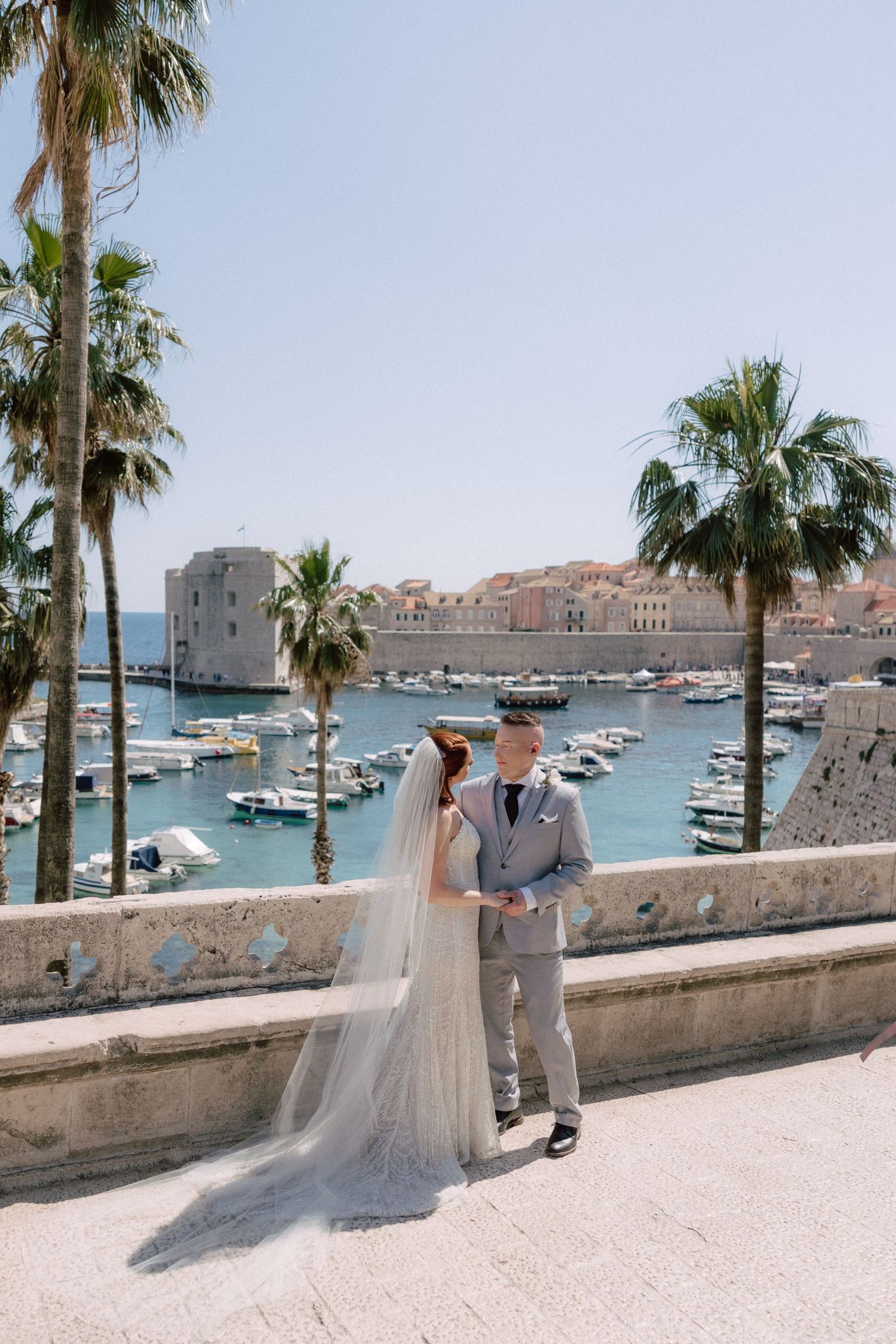 A romantic scene unfolds along Dubrovnik's harbor promenade with palm trees and boats in the background.