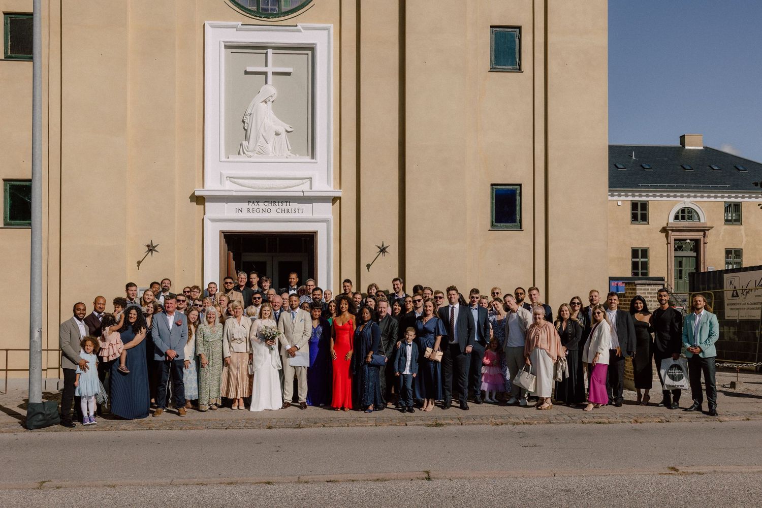 Large group wedding photo of guests gathered outside a church with white cross above entrance.