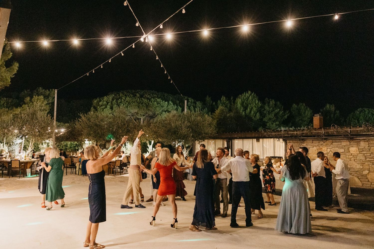 Wedding reception guests dancing under string lights at outdoor venue.