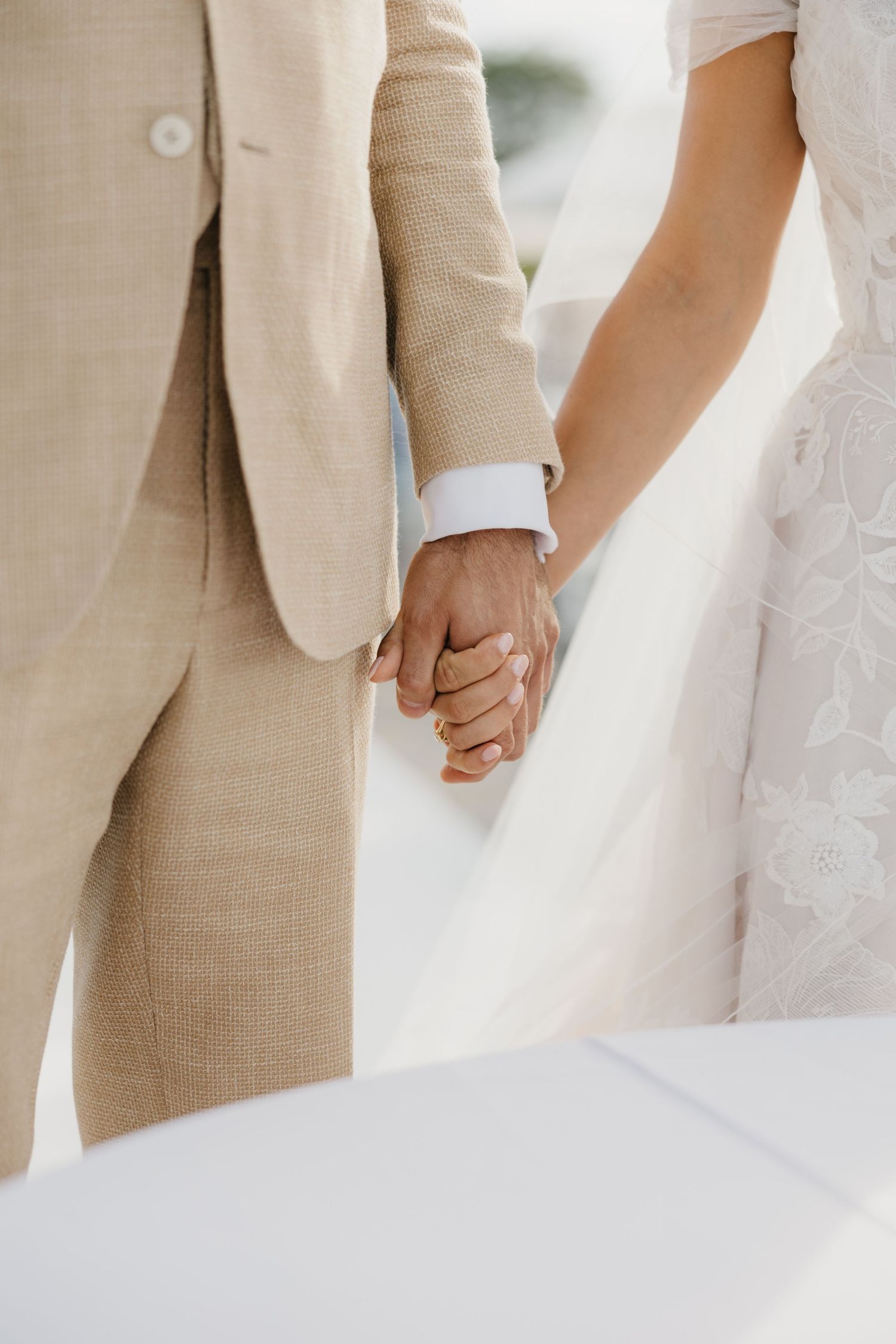 Close-up of couple holding hands, showing beige suit and white dress details.