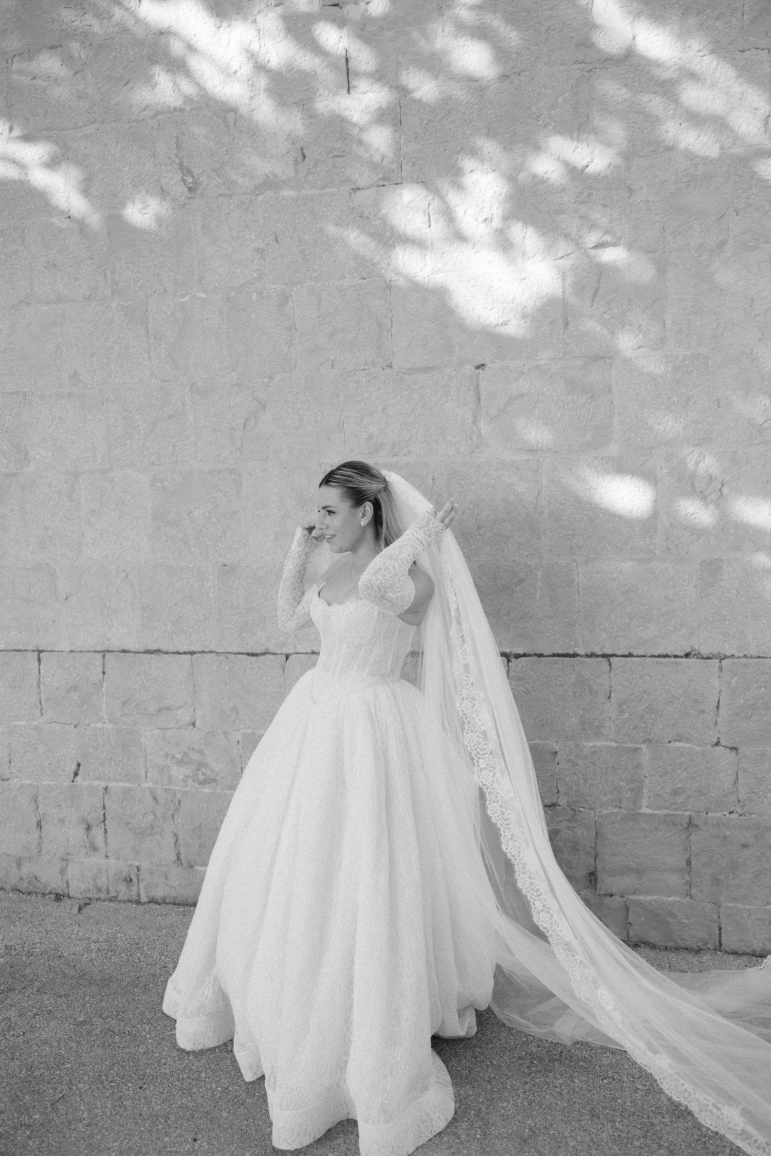 Black and white photo of a bride in a ballgown wedding dress with long lace veil standing against a stone wall.