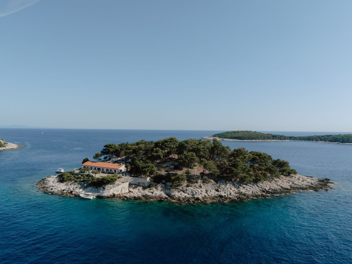 Aerial view of a small rocky island with lush trees and a building surrounded by crystal blue Mediterranean sea.