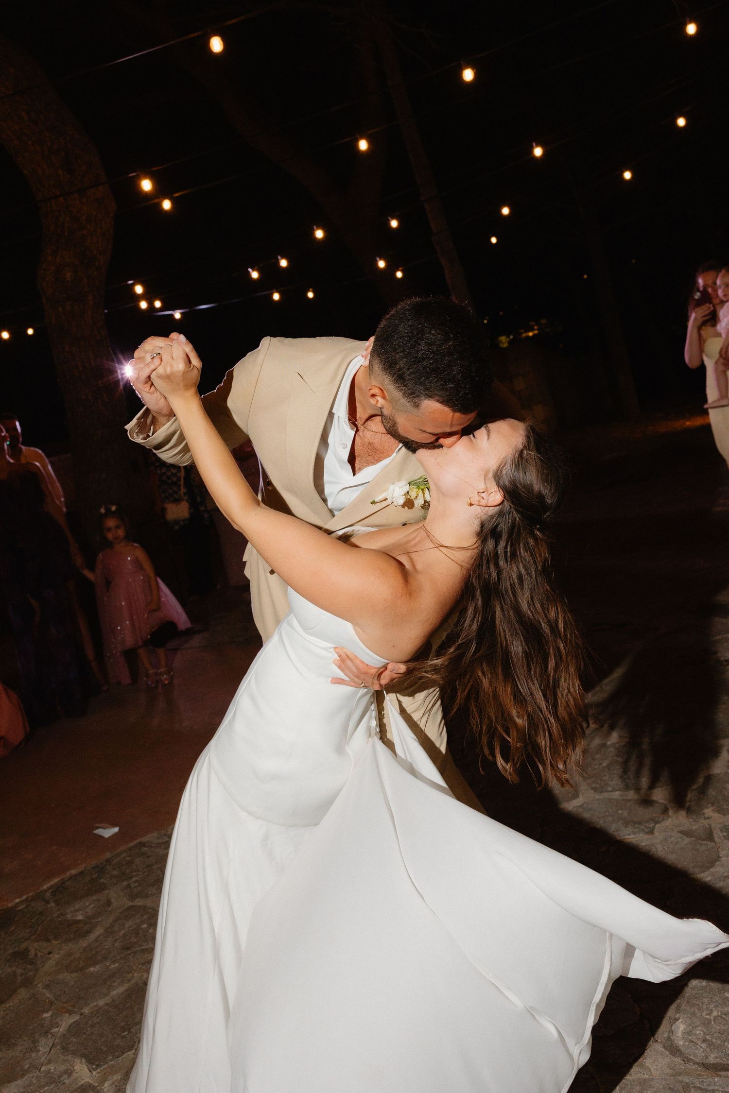 Groom dipping and kissing bride during first dance under string lights at outdoor wedding reception.