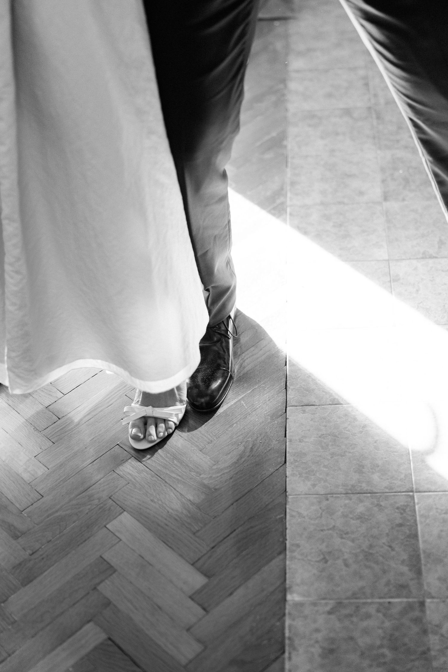 Black and white photo of contrasting formal and casual wedding shoes on a herringbone floor.