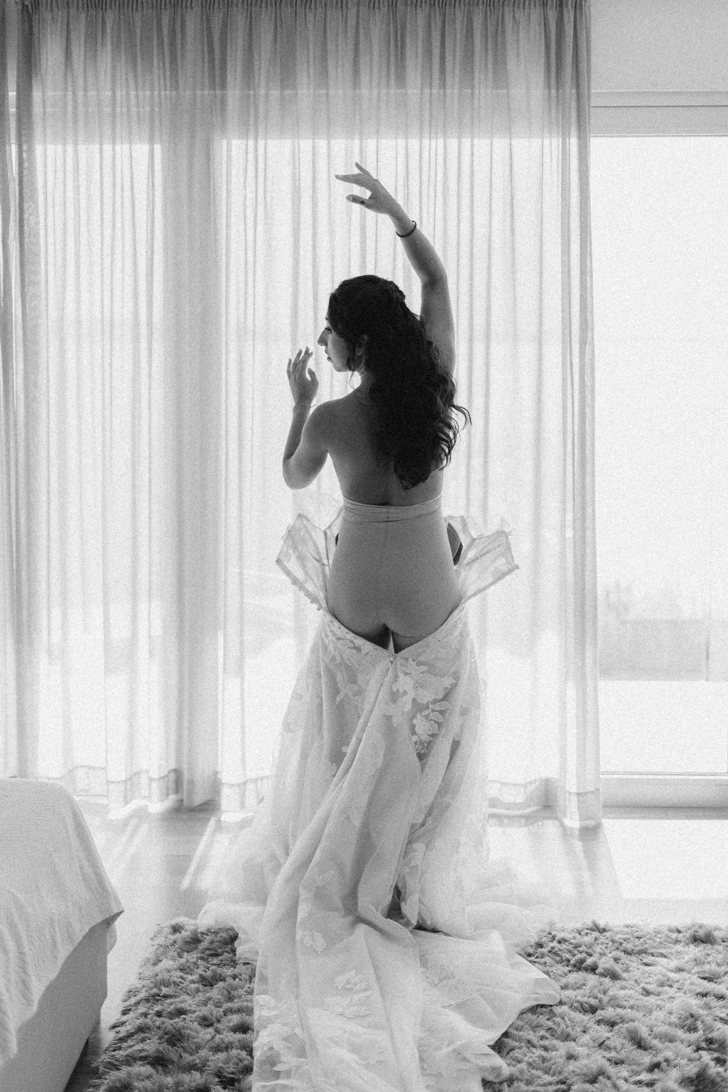 Artistic black and white photo of bride in flowing white dress by window.