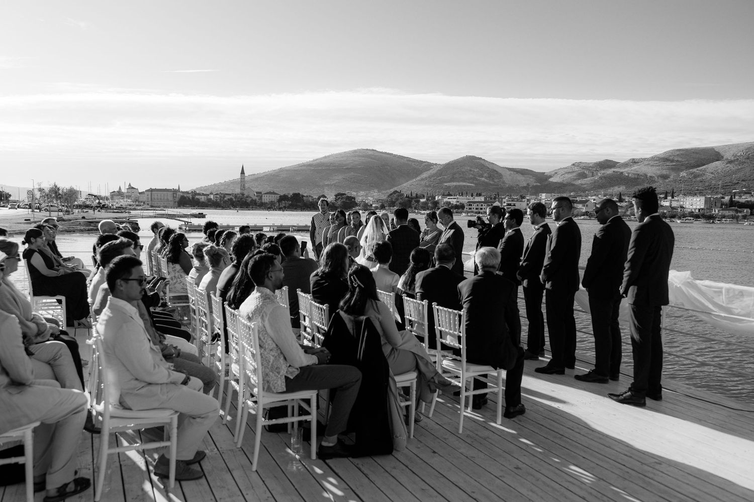 Black and white wide shot of an elegant outdoor waterfront wedding ceremony with guests seated in rows.
