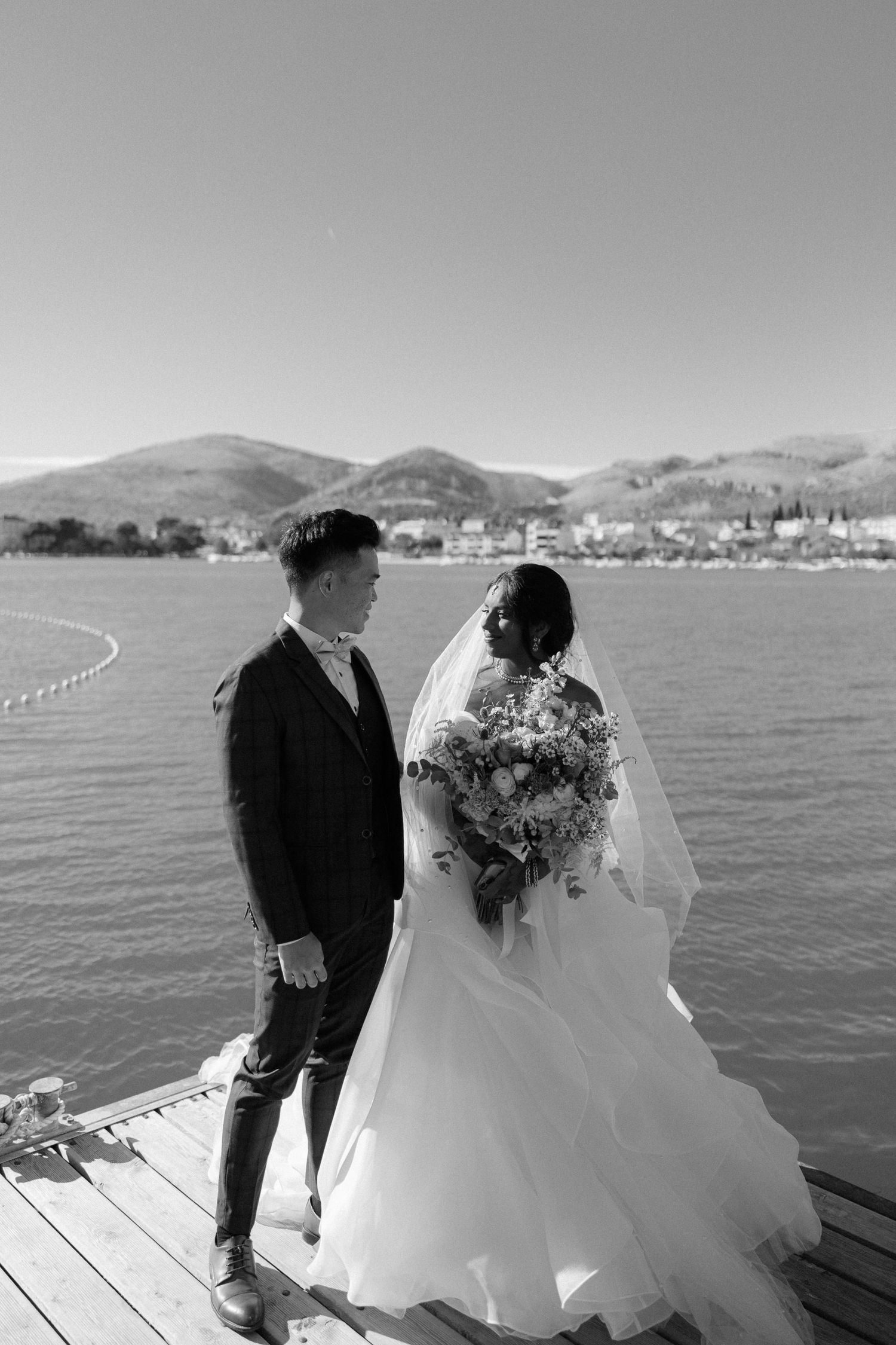 Black and white portrait of bride and groom standing on a wooden dock overlooking a scenic coastal bay.