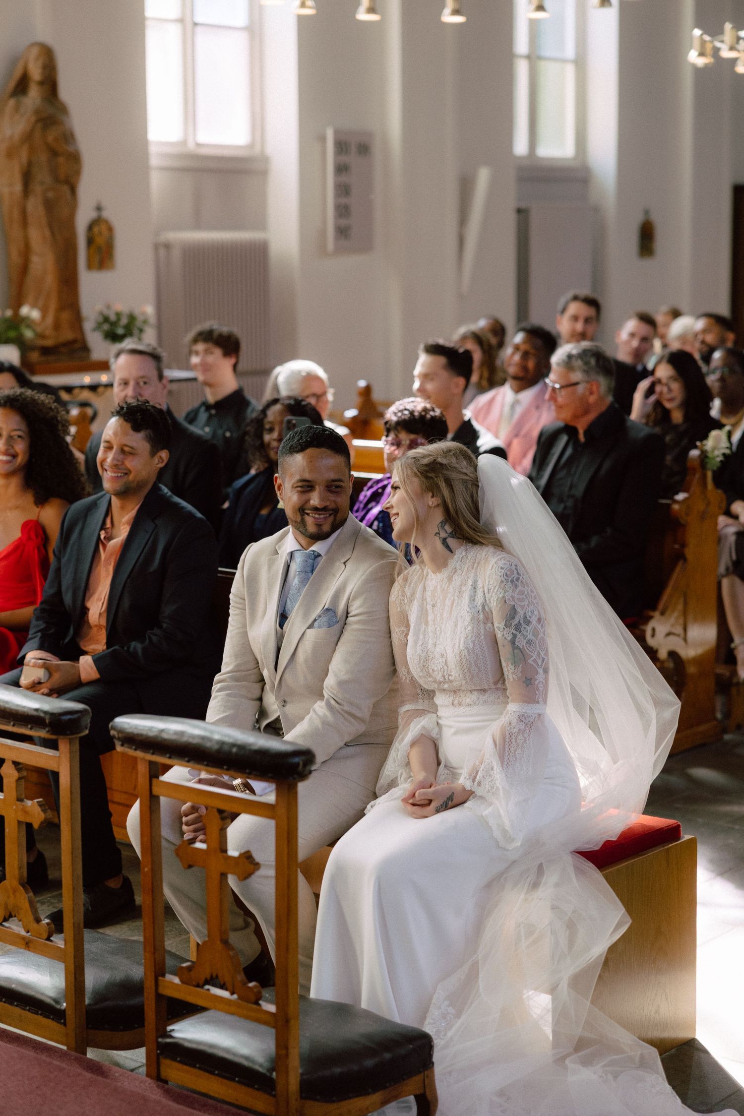 Wedding ceremony attendees seated in wooden church pews with decorative lighting and religious statues.