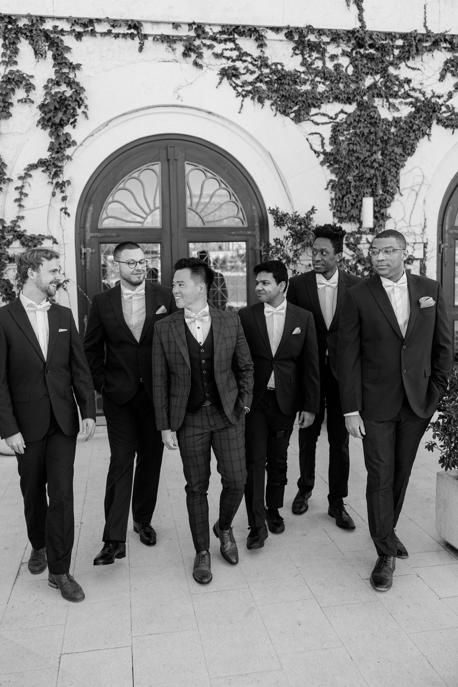 Groom in plaid suit walks with groomsmen in black suits outside ivy-covered venue.