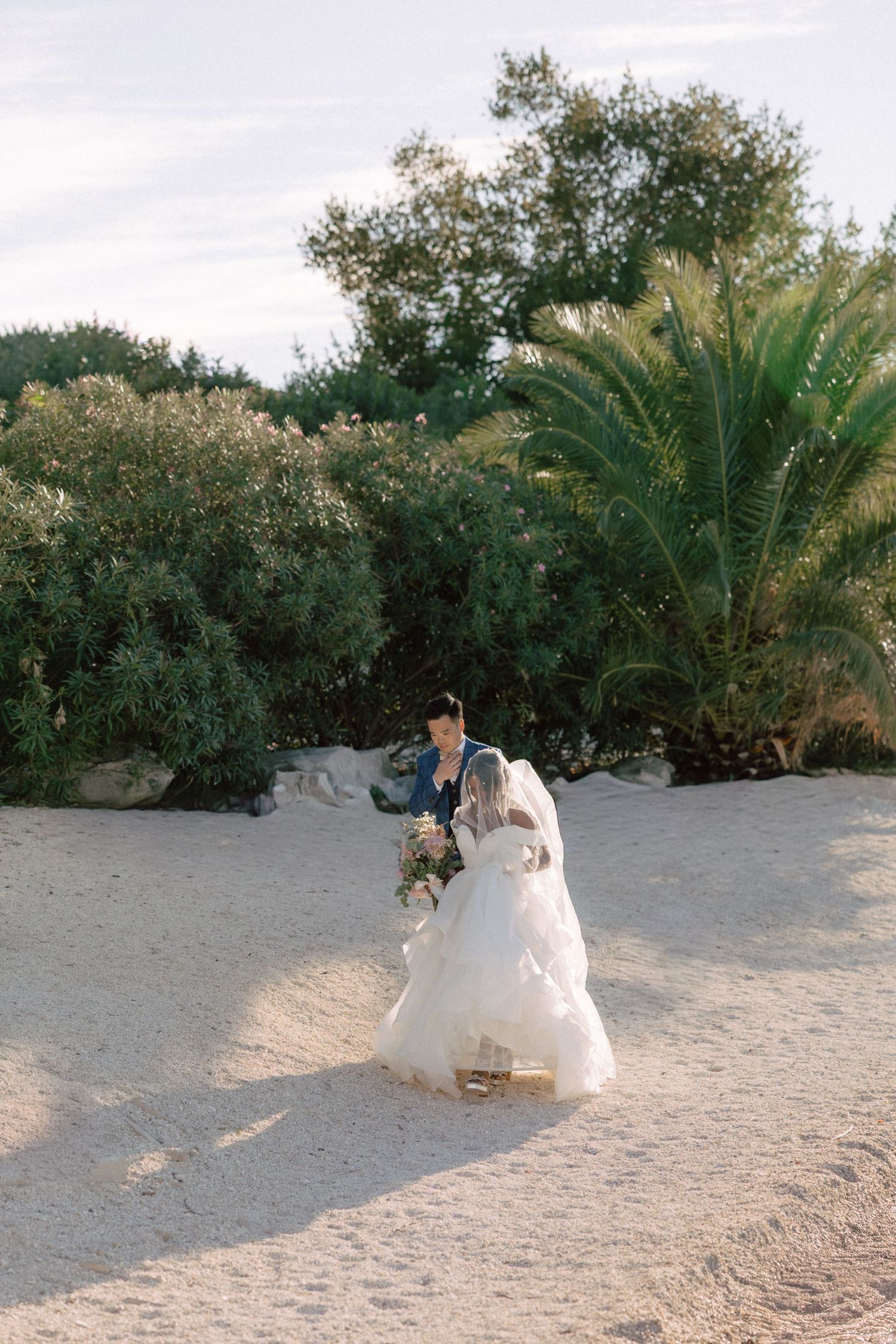 Bride and groom walking together on sandy beach surrounded by lush green trees and palm.