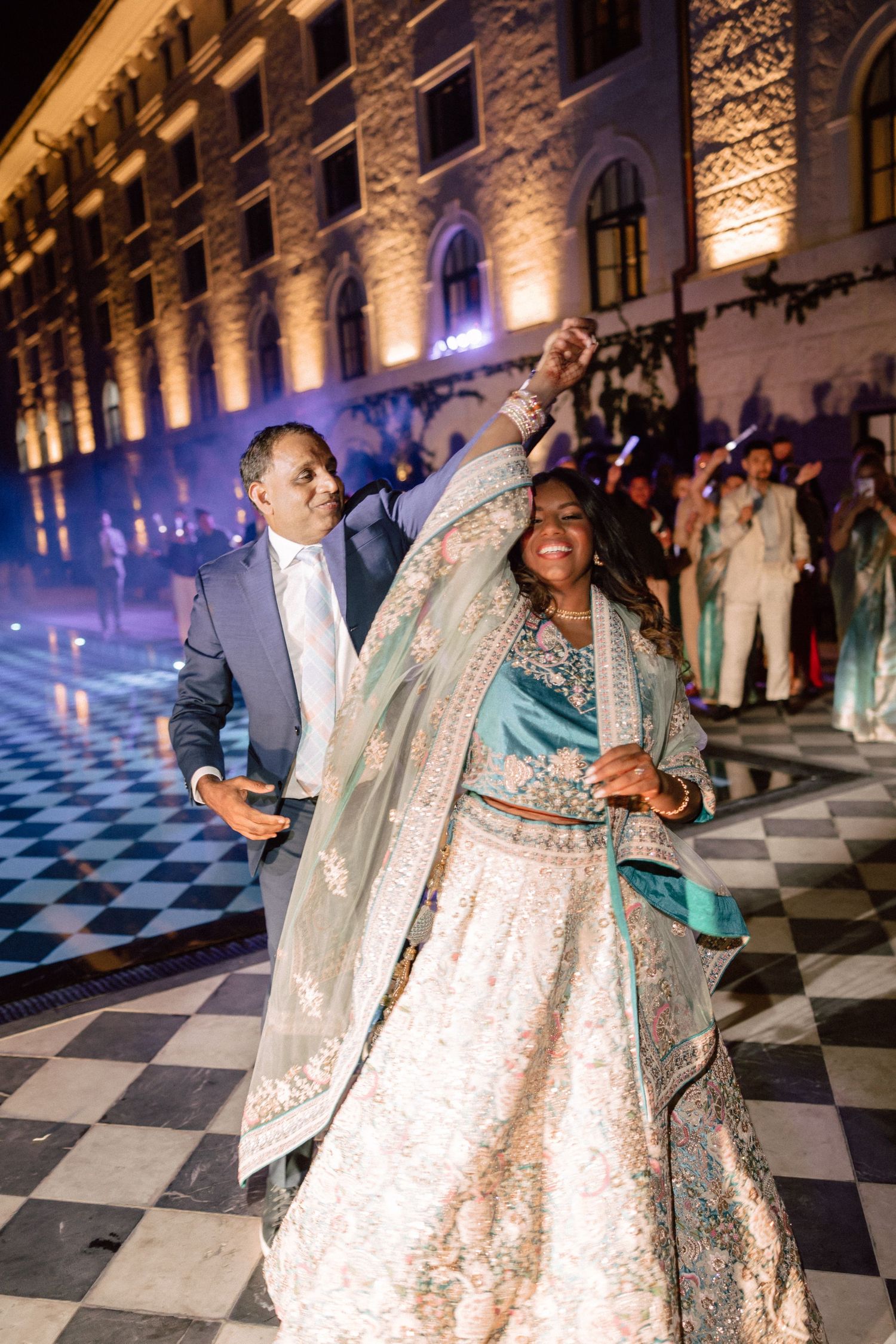 Bride in teal and gold lehenga dancing with partner on checkered floor at night wedding reception.