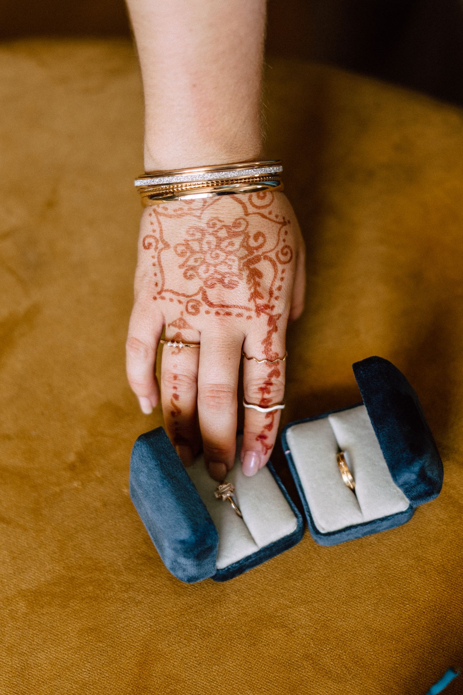 Bride's hand adorned with intricate henna mehndi design, gold bangles, and rings beside two wedding ring boxes.