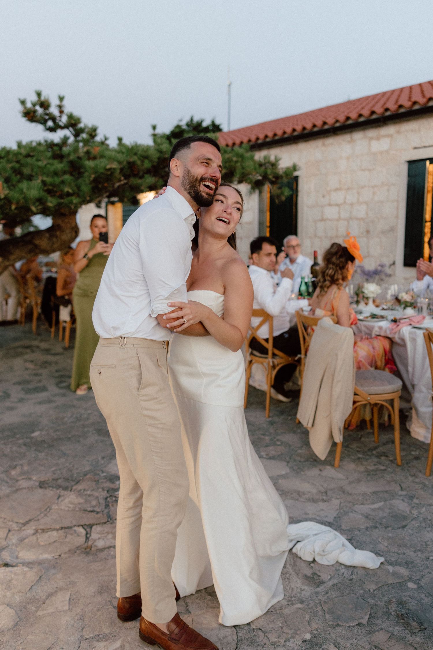 Happy bride and groom embracing and laughing outdoors at their Mediterranean island wedding reception at sunset.