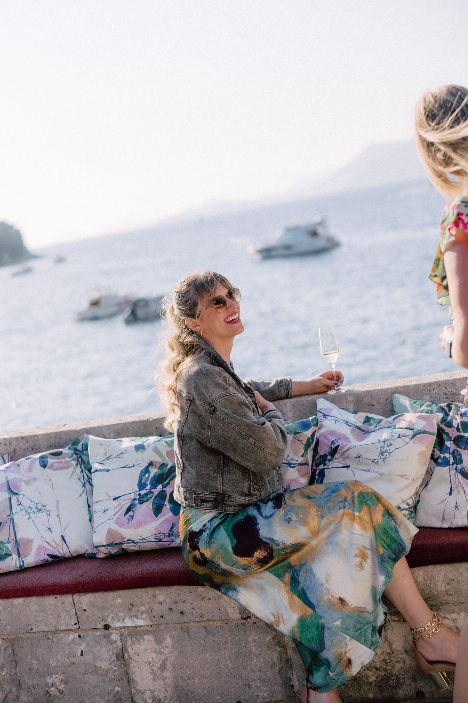 People relaxing on seaside wall with floral cushions and boats visible in background.