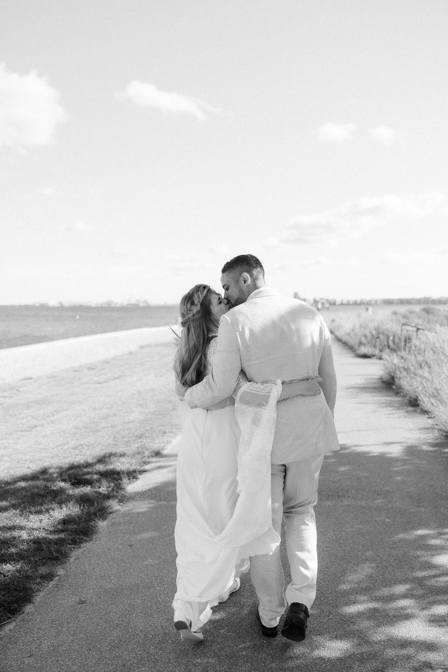 A couple in white wedding attire walks away from camera along a scenic coastal path.