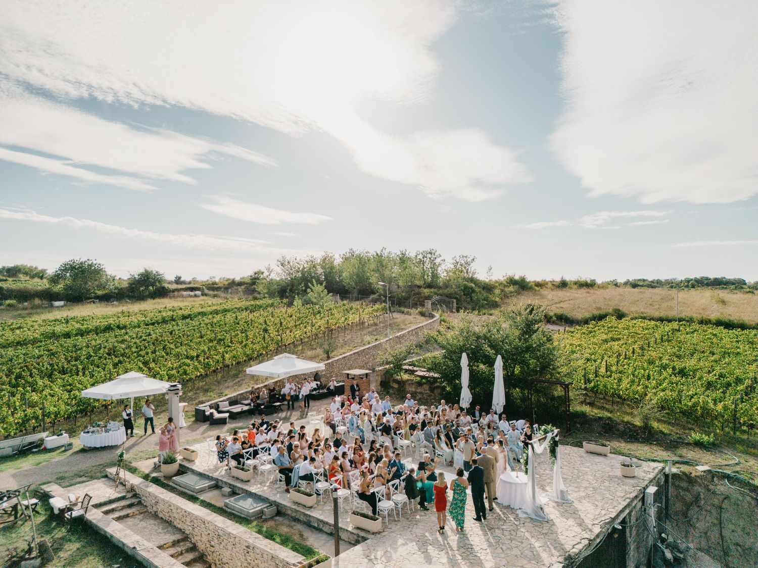 Aerial view of outdoor wedding reception with guests gathered on stone patio surrounded by vineyard in Italy.