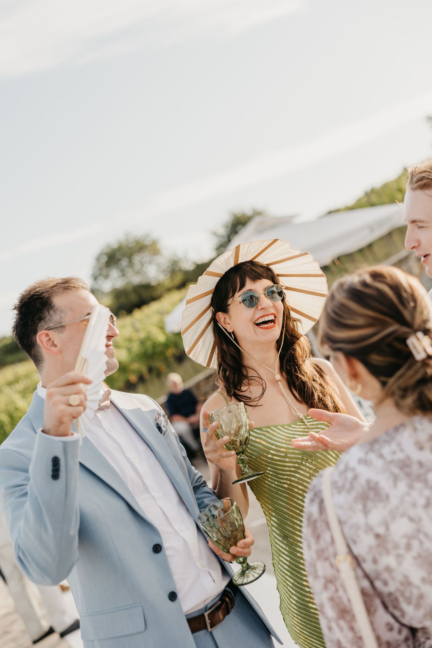 Wedding party laughs together under sunlight while holding decorative paper parasols.
