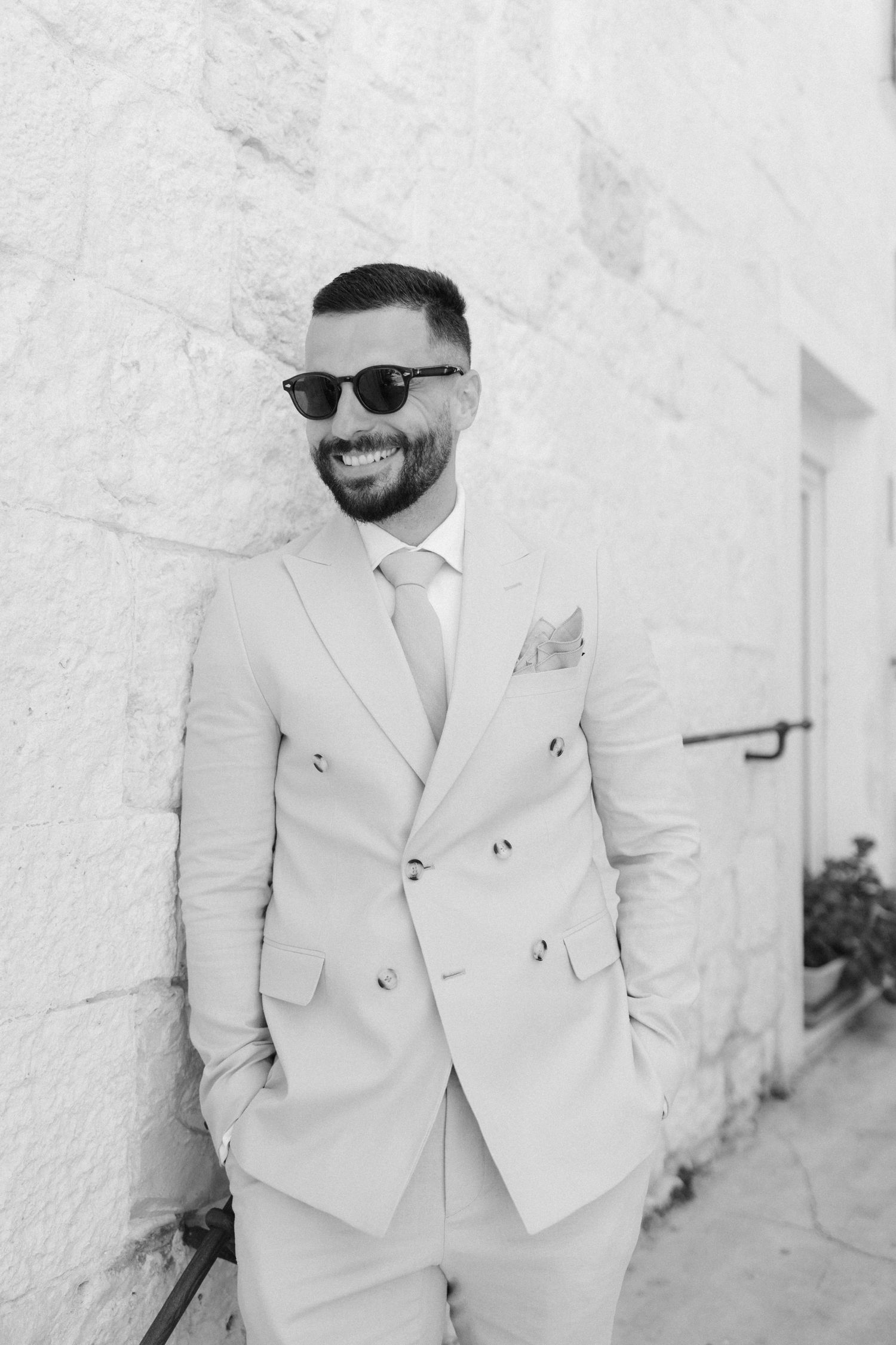 Stylish groom in cream double-breasted suit and sunglasses smiling against white stone wall.