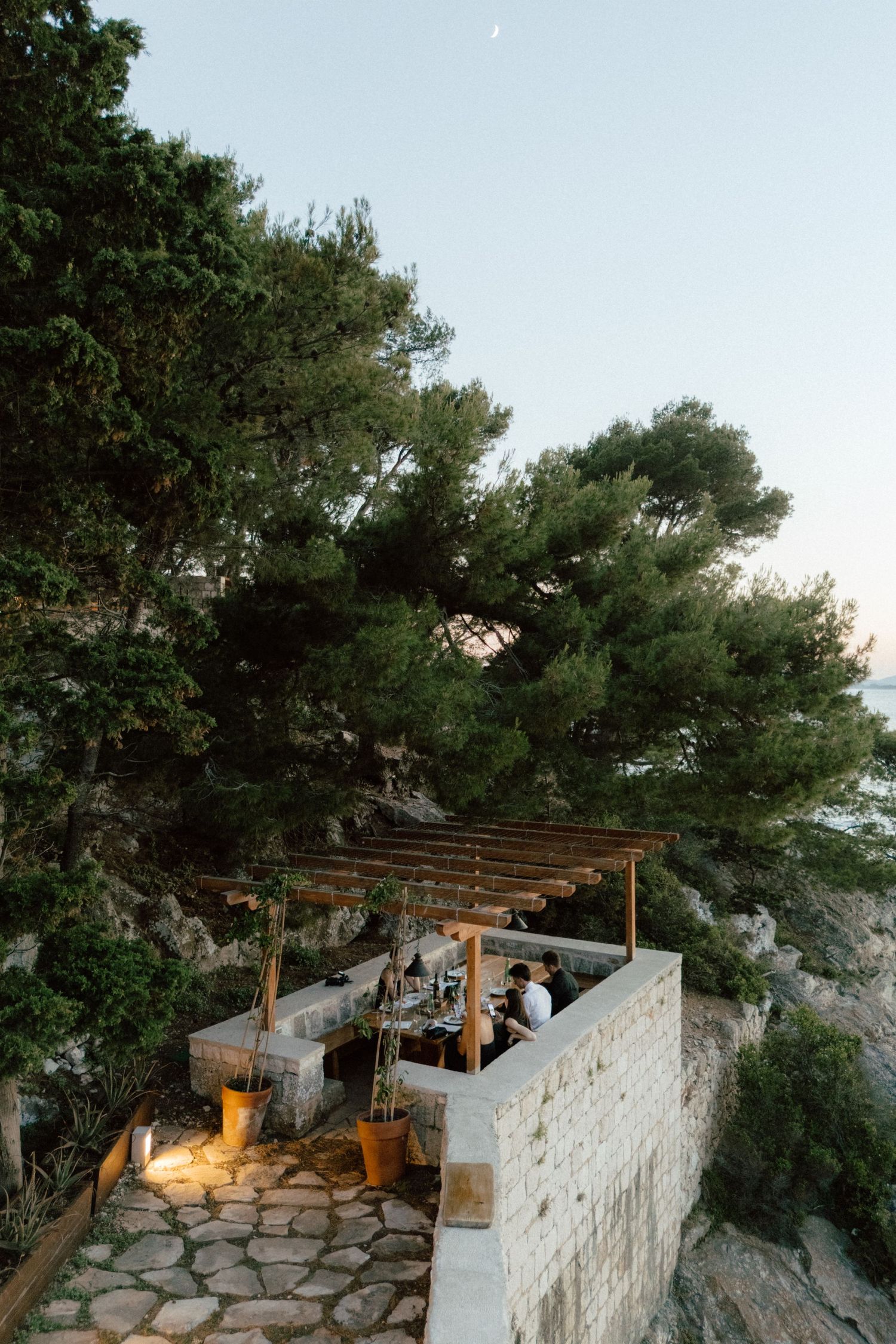Romantic cliffside pergola dining area overlooking the sea at dusk, surrounded by lush pine trees on rocky Mediterranean coast.
