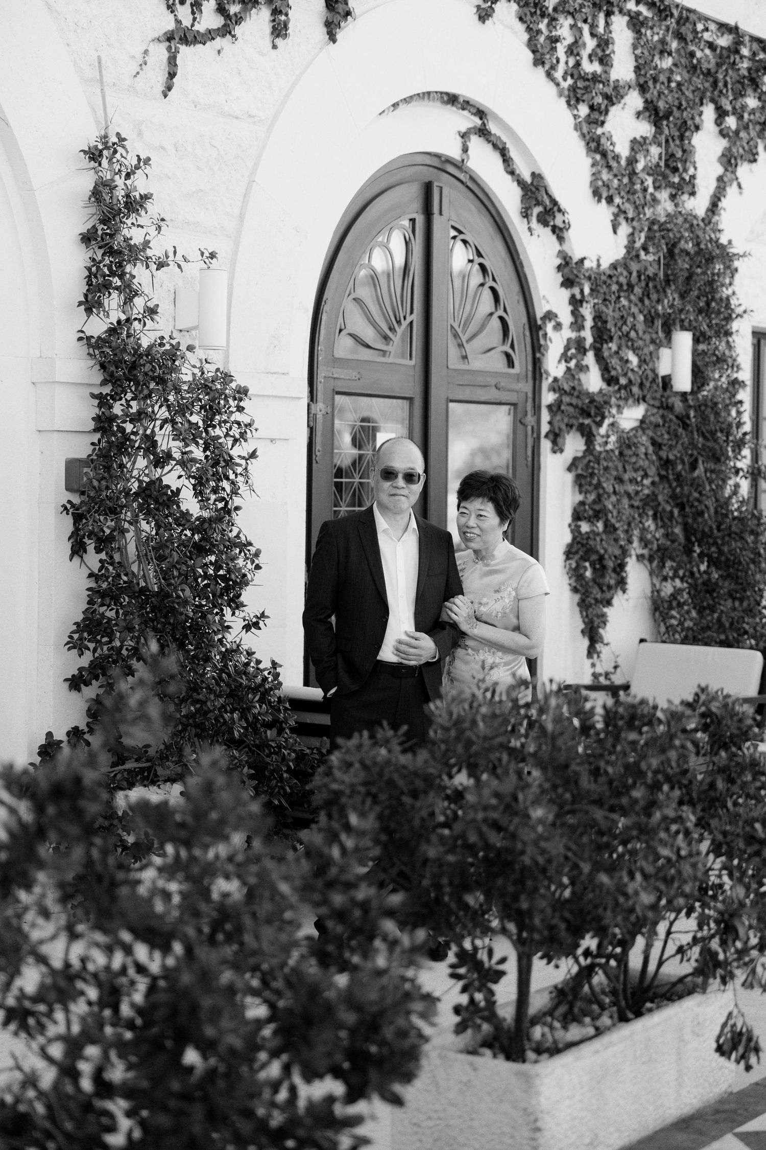 Elegant couple poses outside ivy-covered white building with arched wooden door in black and white photo.