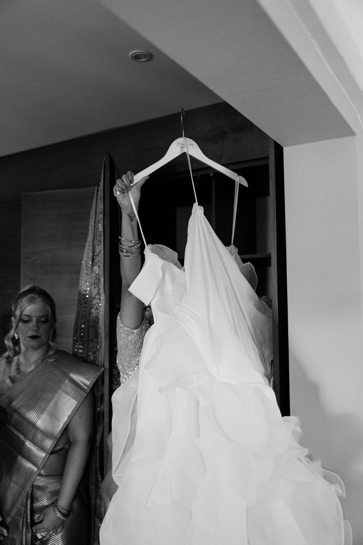 Black and white photo of a ruffled wedding dress on a hanger held up in a hotel room doorway.
