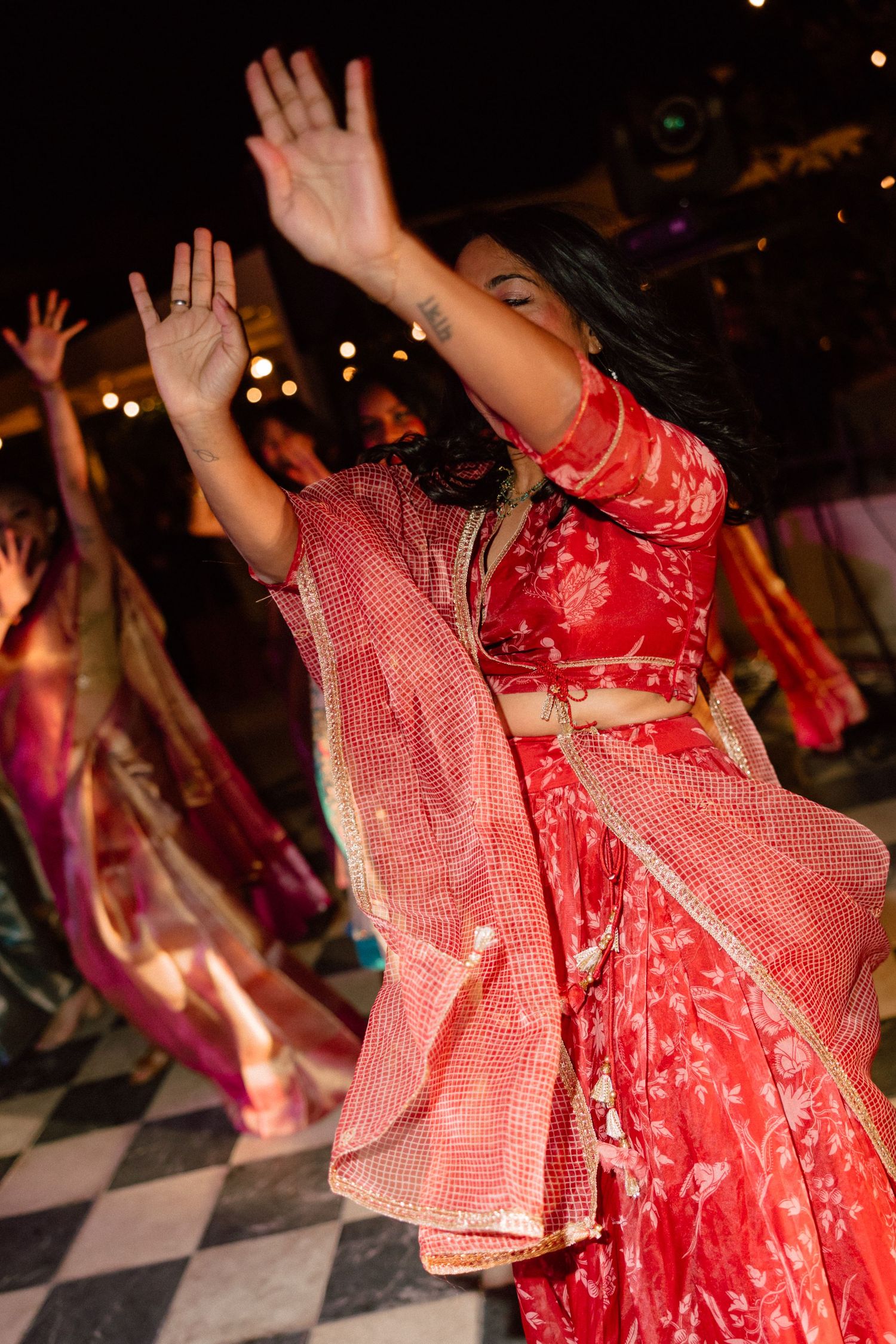 Woman in red lehenga dancing gracefully at an Indian wedding celebration with other guests on a checkered dance floor.