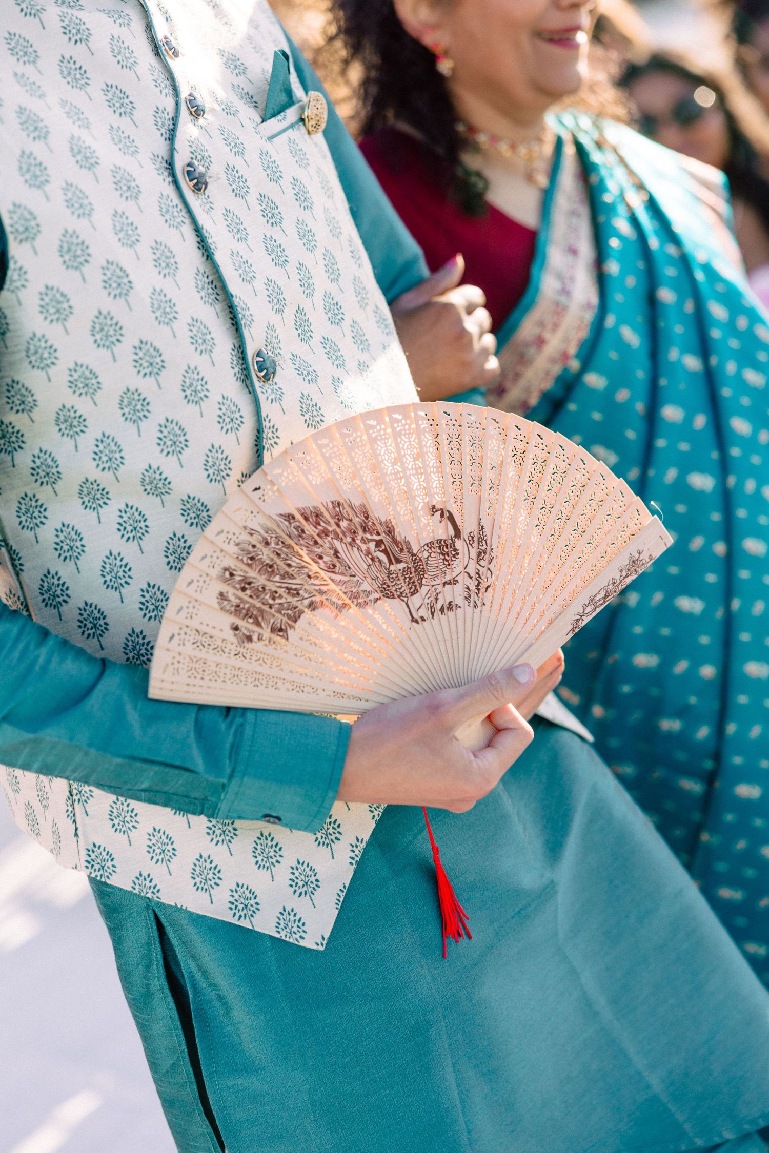 Guest in teal Indian attire holding an ornate wooden hand fan with red tassel at outdoor wedding.