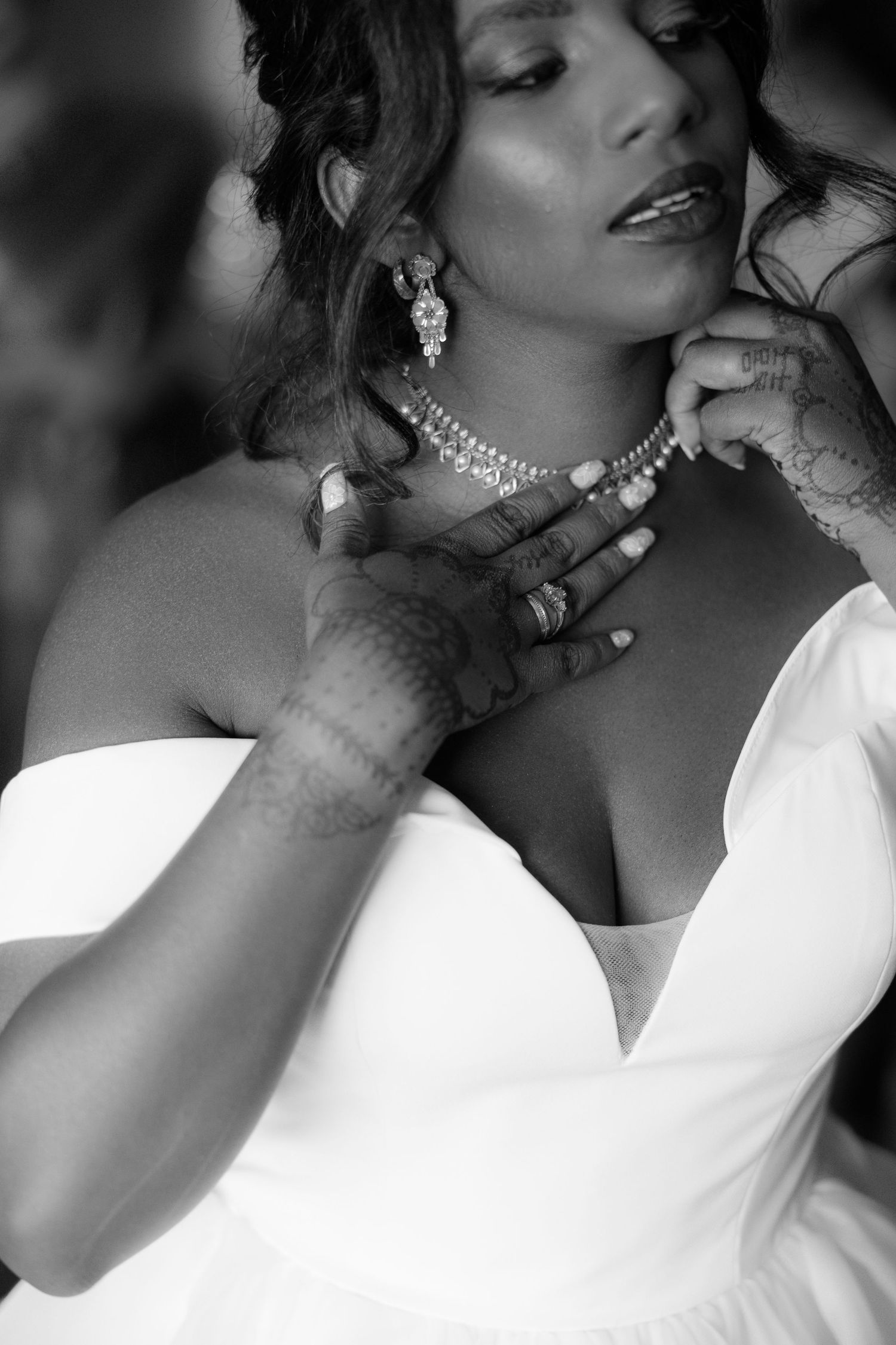 Black and white portrait of bride in white gown adjusting diamond necklace with henna hands.