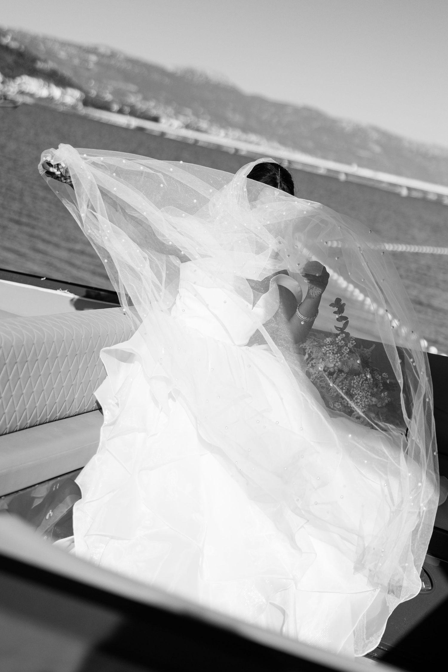 Bride in flowing white gown and veil stepping out of a car with scenic waterfront in black and white.