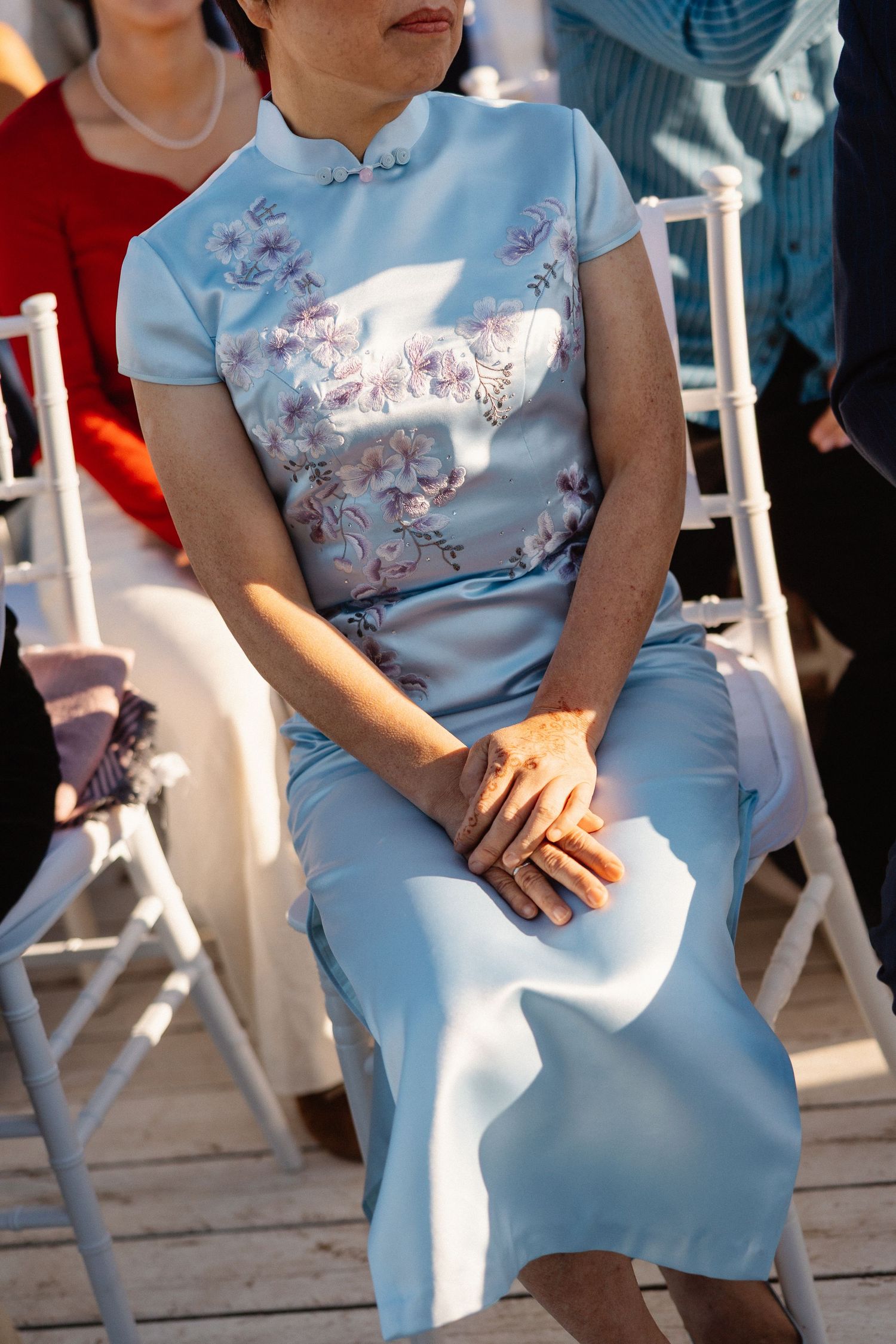 Elegant older woman in light blue floral cheongsam dress seated at an outdoor wedding ceremony.
