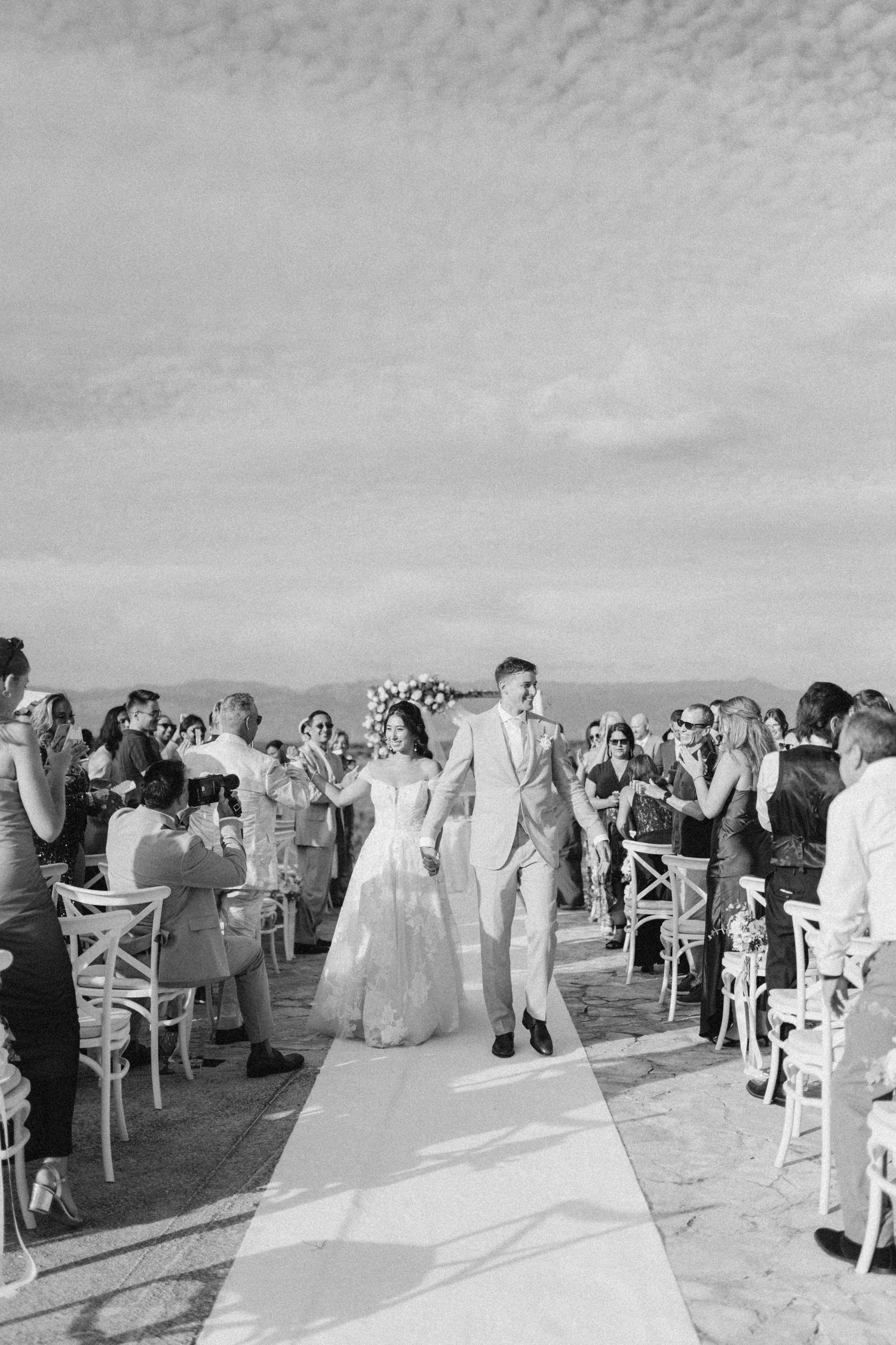 Black and white panorama of wedding ceremony guests gathered outdoors under cloudy sky.