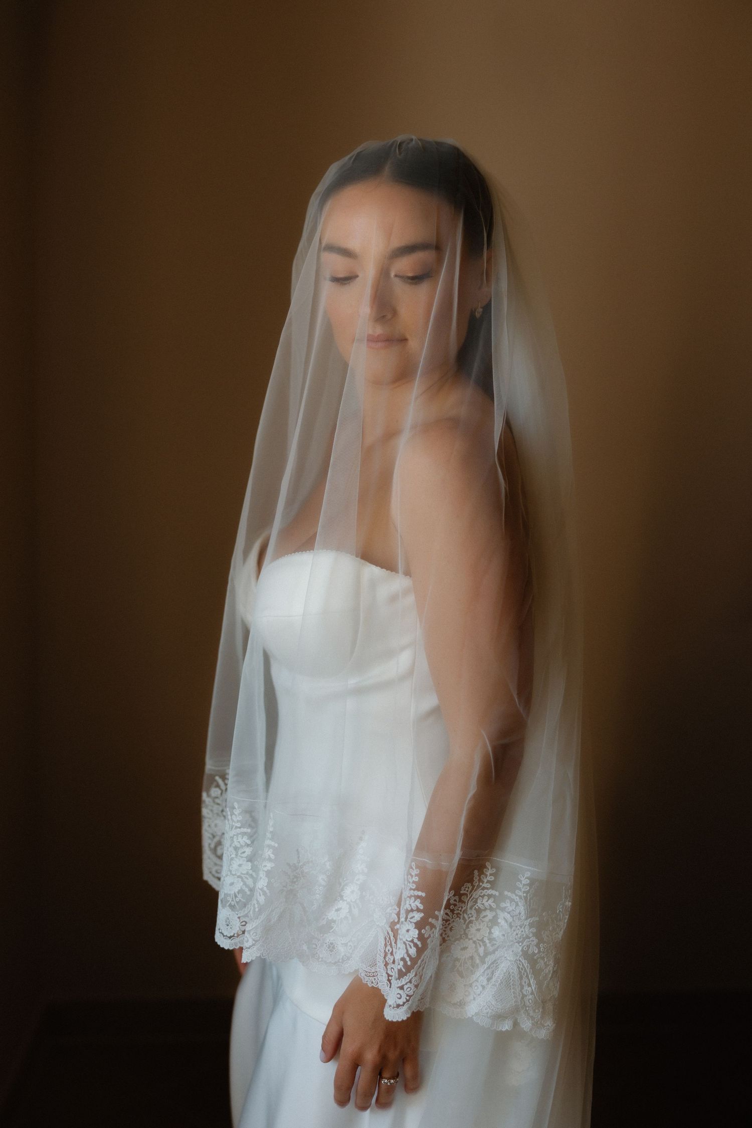 Bride in white strapless gown and lace-trimmed veil with eyes closed, posing elegantly against a warm brown background.