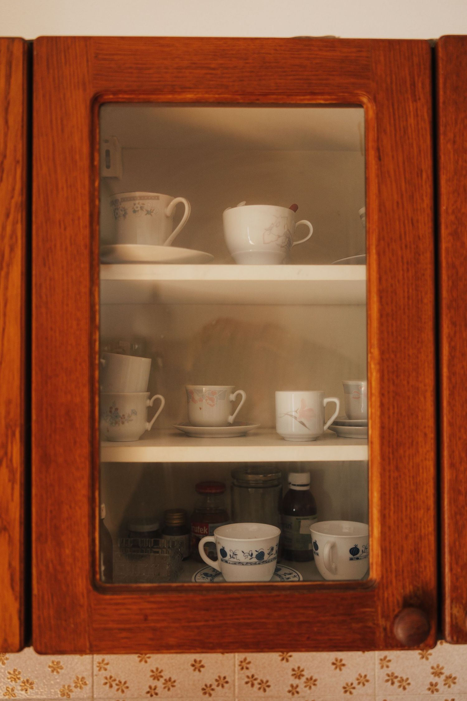 Vintage wooden kitchen cabinets with glass doors displaying white teacups and mugs on shelves.