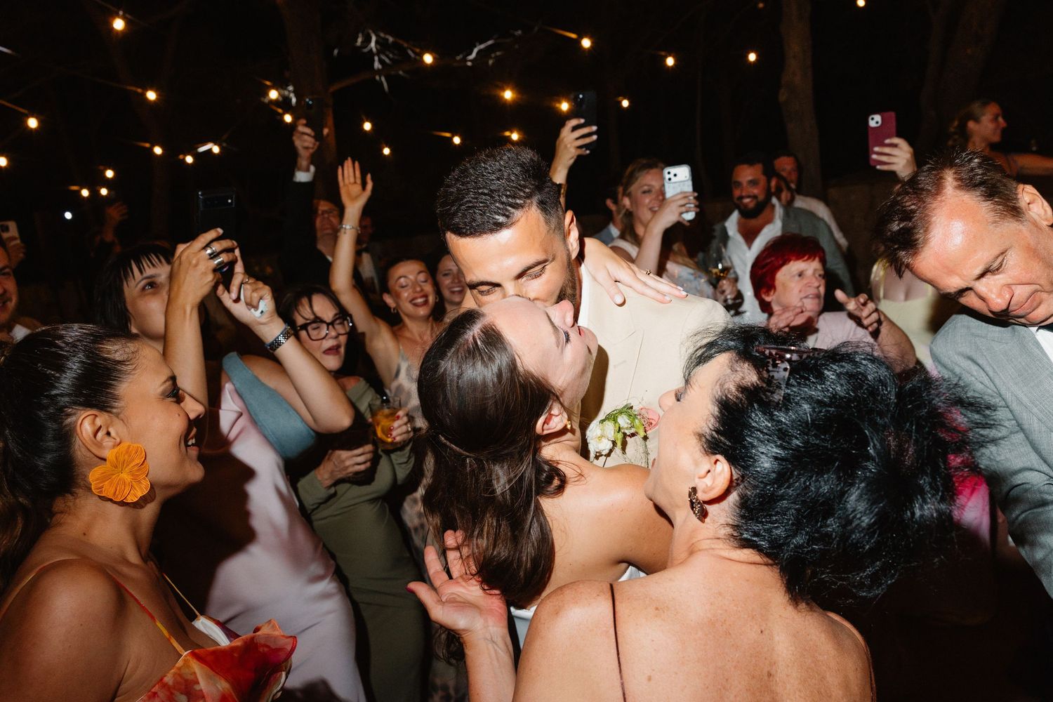 Bride and groom share a romantic kiss surrounded by cheering wedding guests at a lively outdoor reception at night.