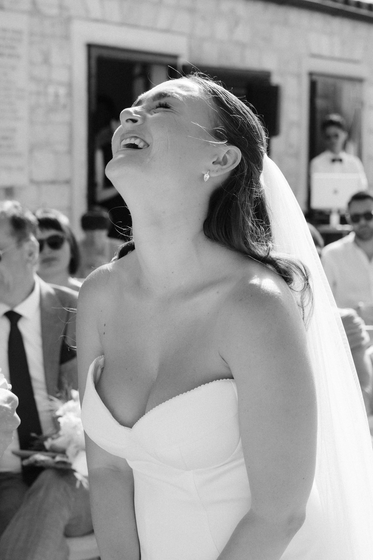 Black and white photo of a joyful bride in a strapless gown laughing with head tilted back at outdoor wedding.