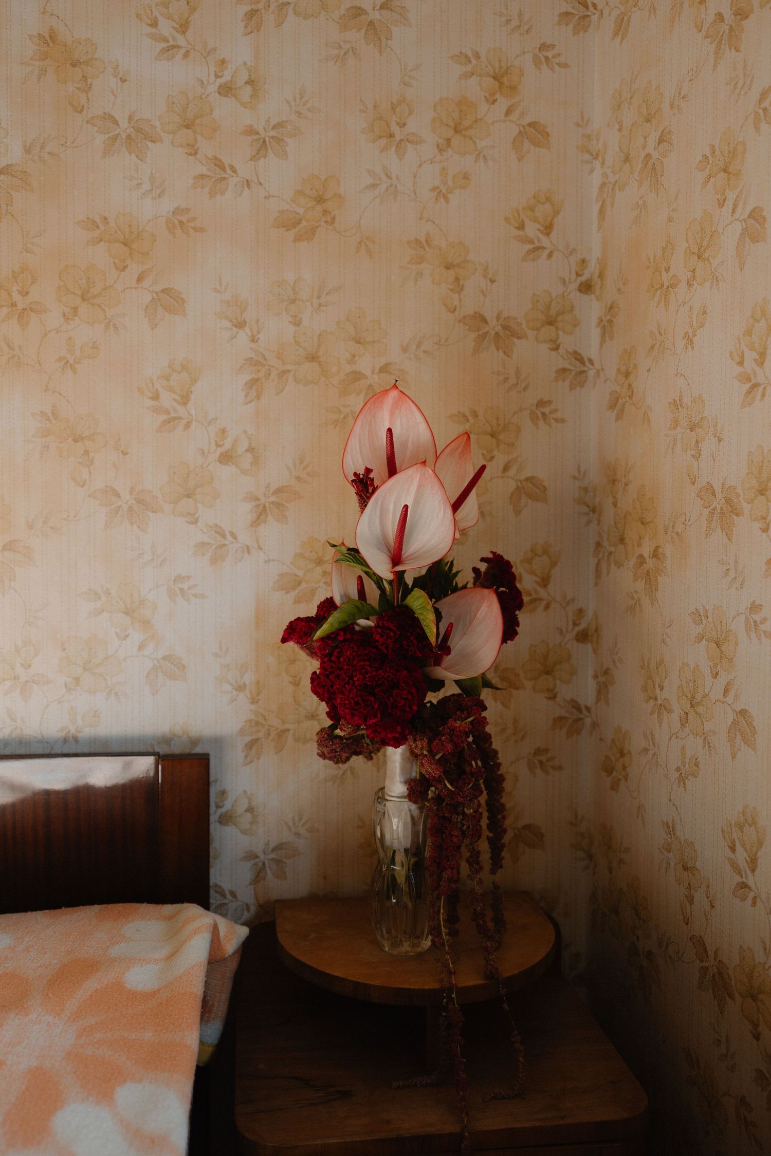 Pink tulips and deep red flowers in a glass vase on wooden side table next to bed with floral wallpaper background.