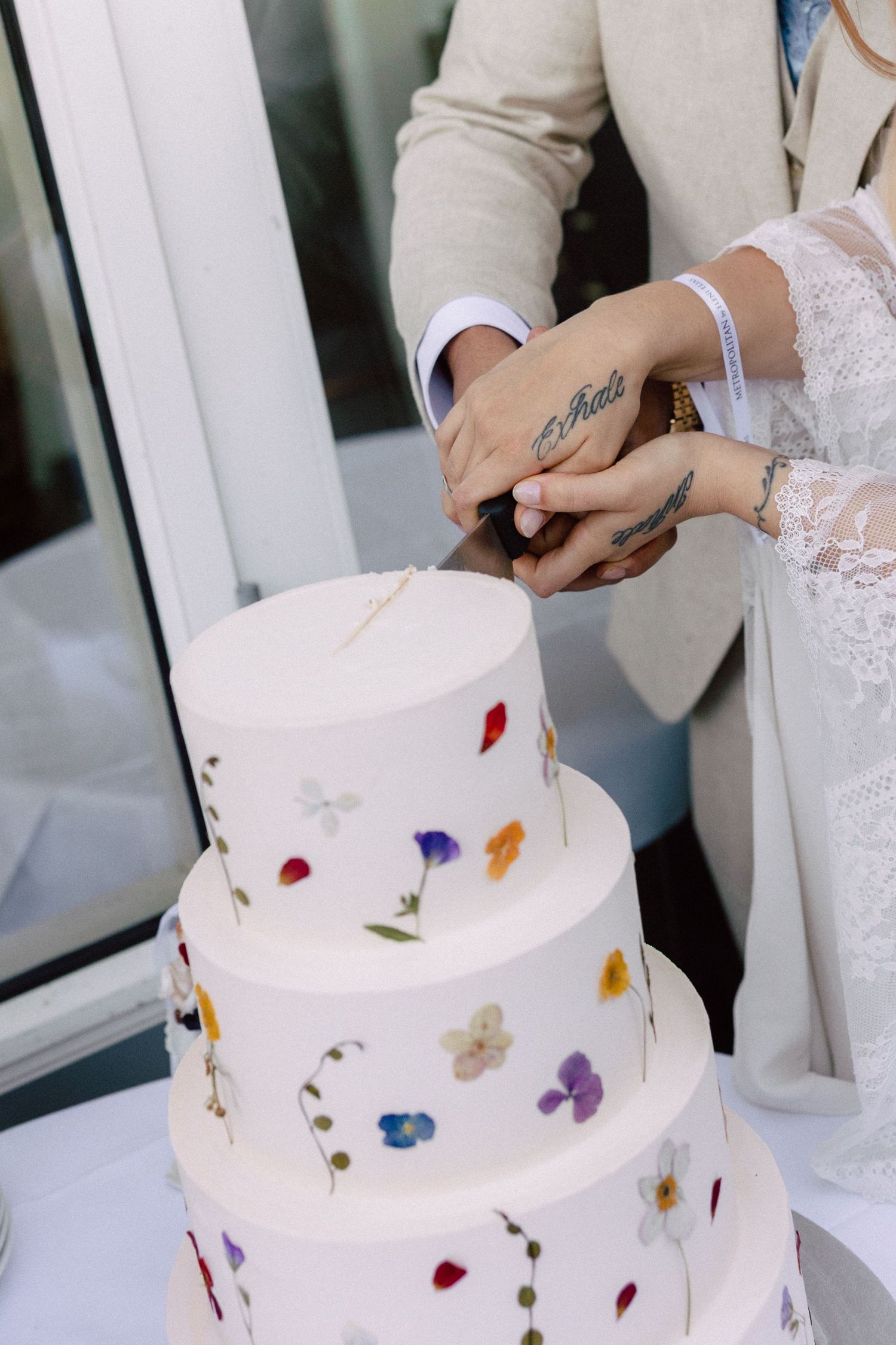 Couple cuts into their white wedding cake decorated with colorful pressed flowers.