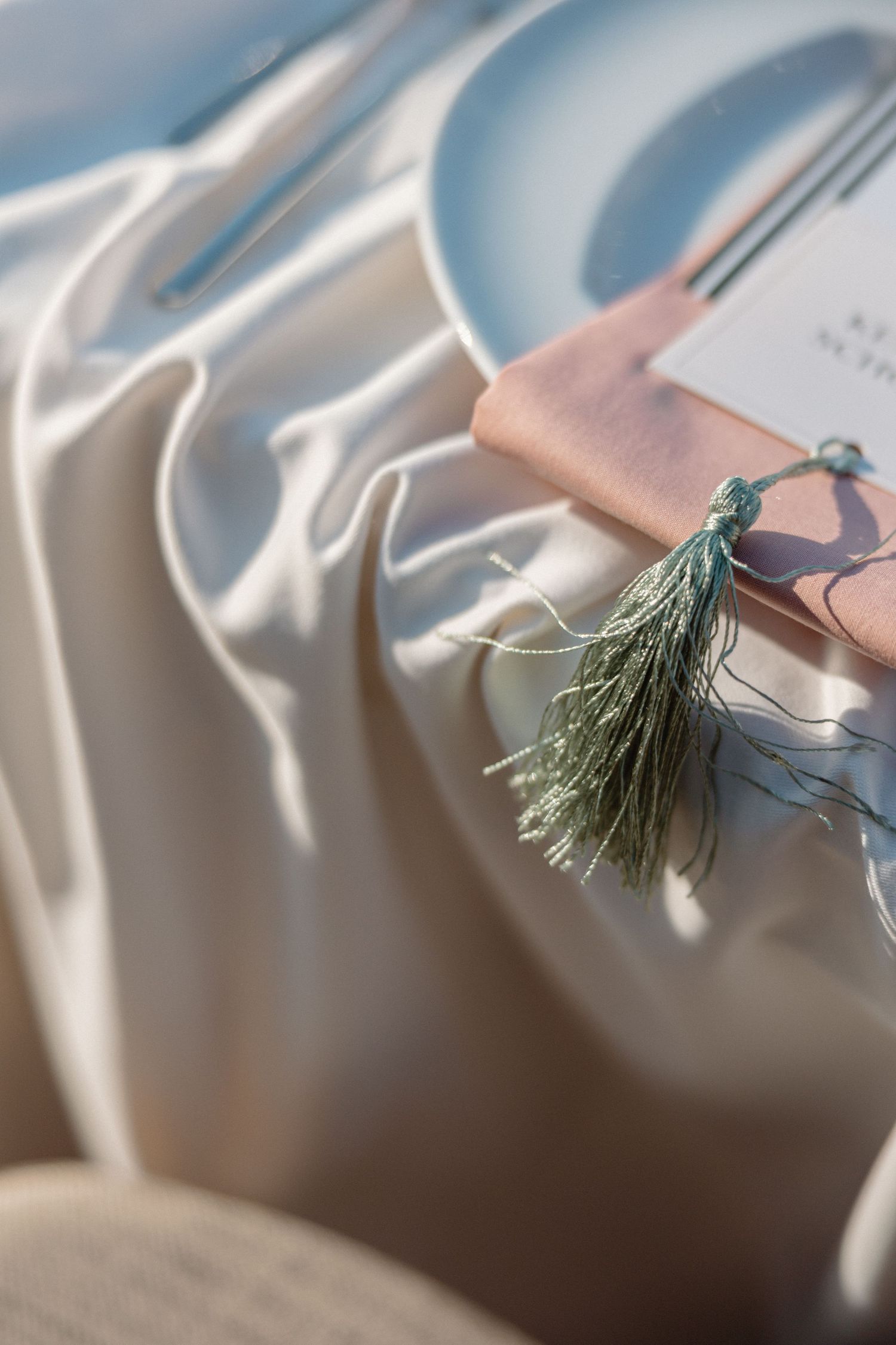 Elegant wedding place setting with cream linen, white plate, and teal tassel gift in sunlight.