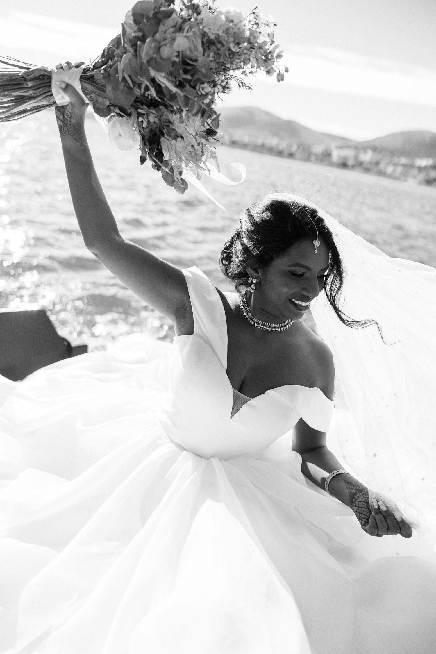 Black and white photo of joyful bride in ball gown raising bouquet near water.