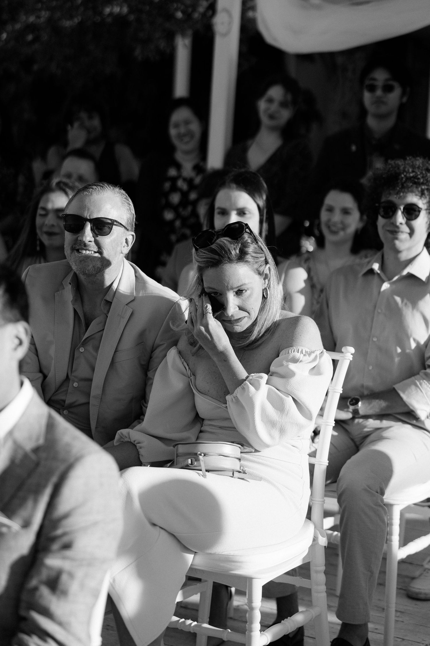 Emotional wedding guest wipes tears while seated among smiling attendees at outdoor ceremony.