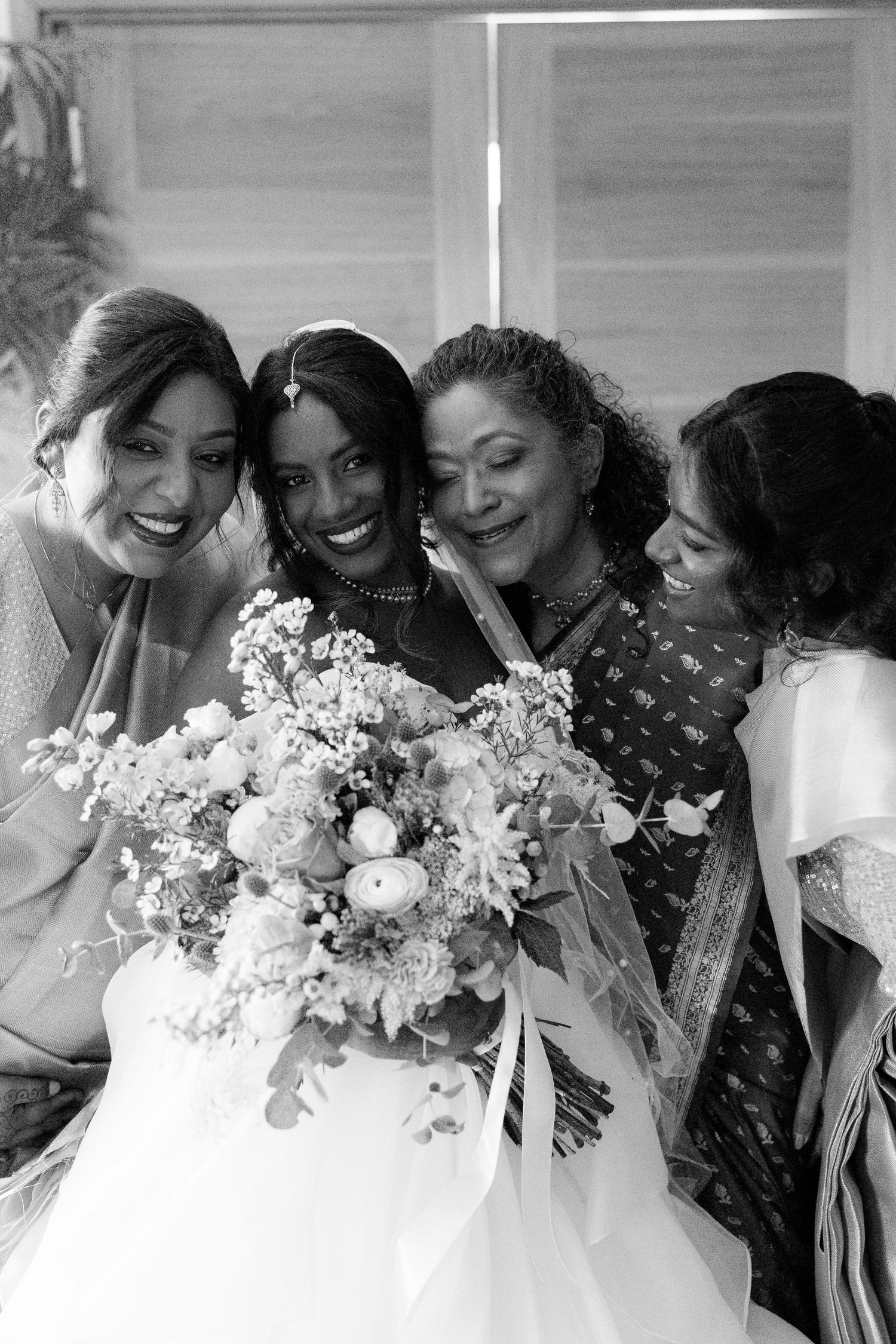 Black and white photo of a radiant South Asian bride with bouquet sharing a joyful moment with three women.