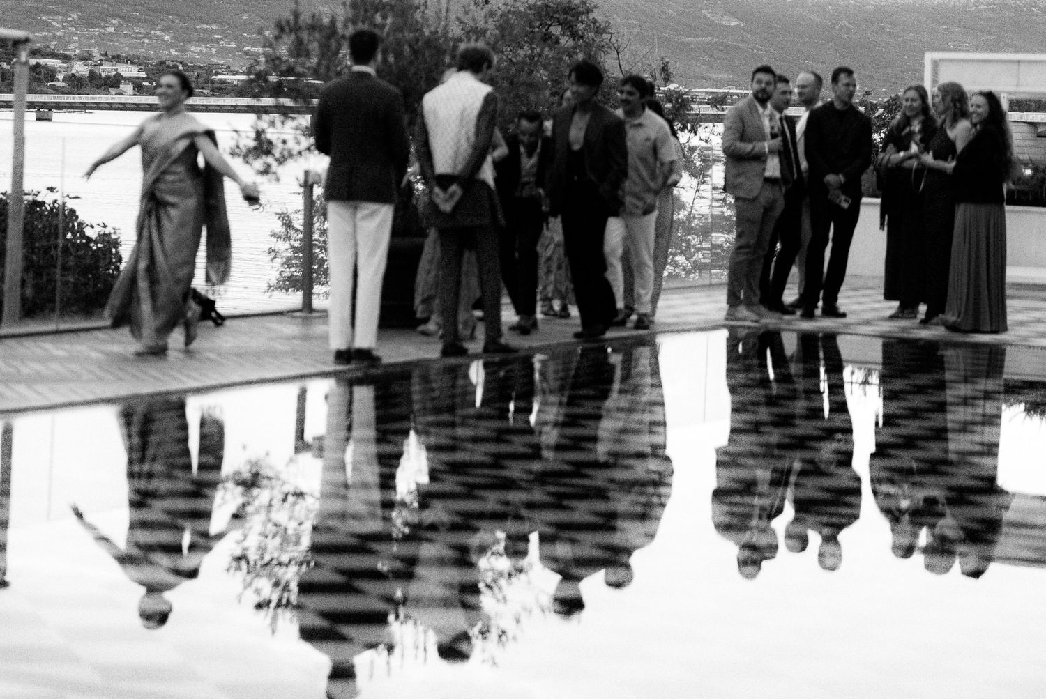 Wedding guests mingle poolside with reflections visible in water, black and white.