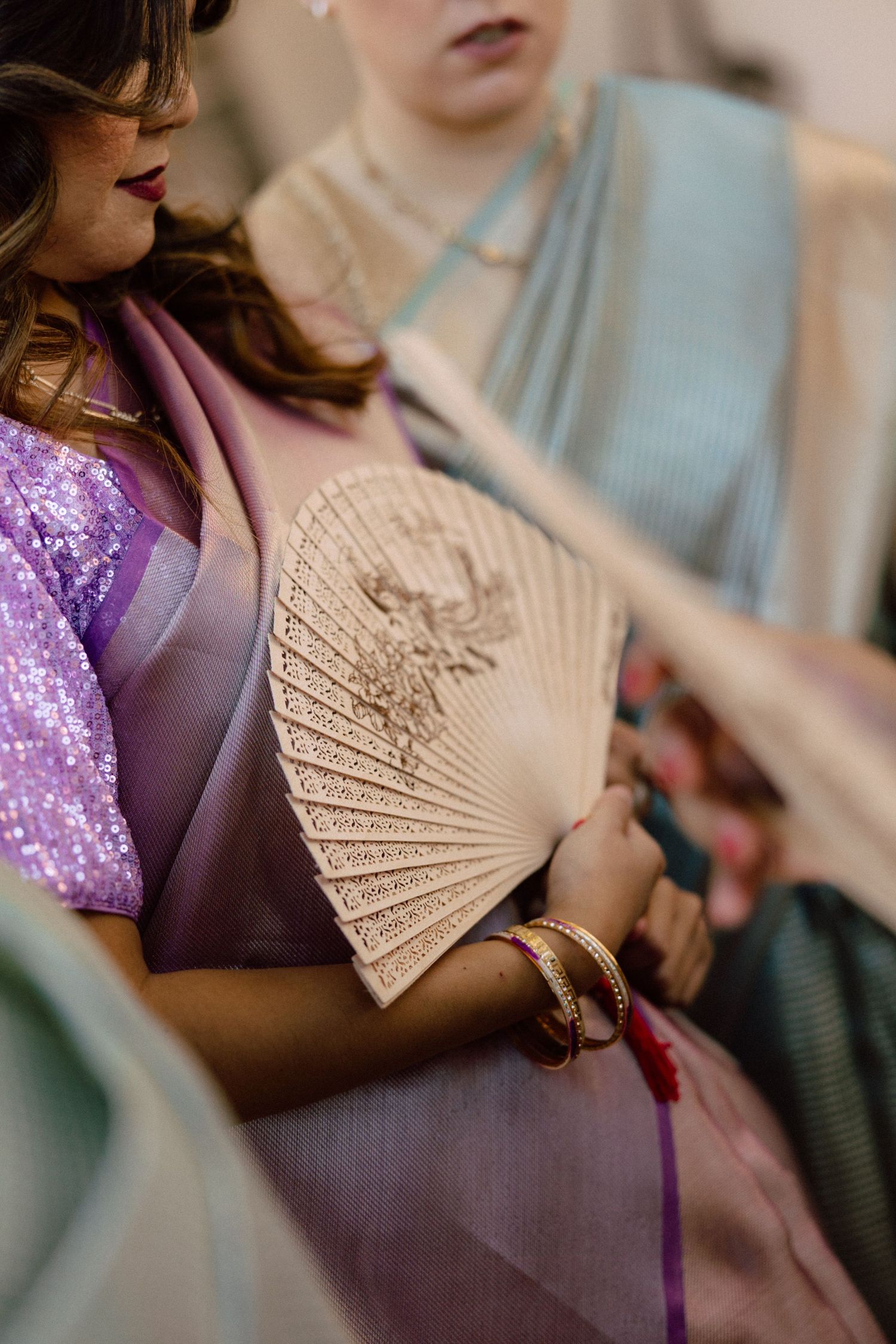 Woman in lavender saree holding an ornate carved fan with gold bangles at an Indian wedding celebration.