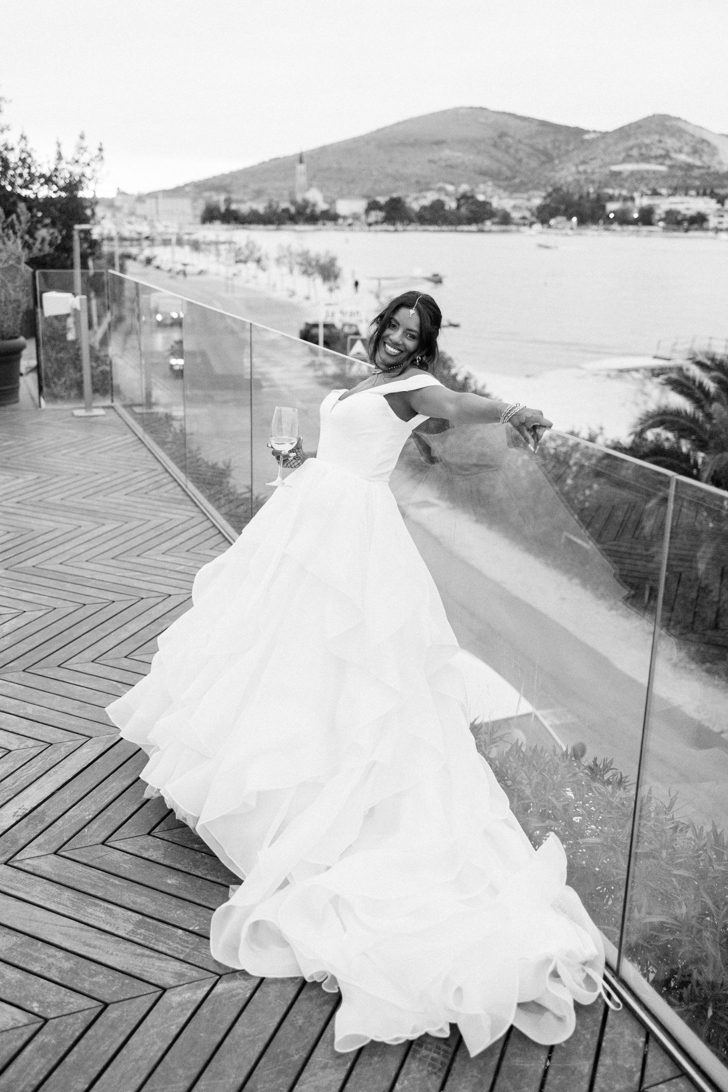 Black and white portrait of bride in voluminous white wedding gown on glass balcony overlooking water.