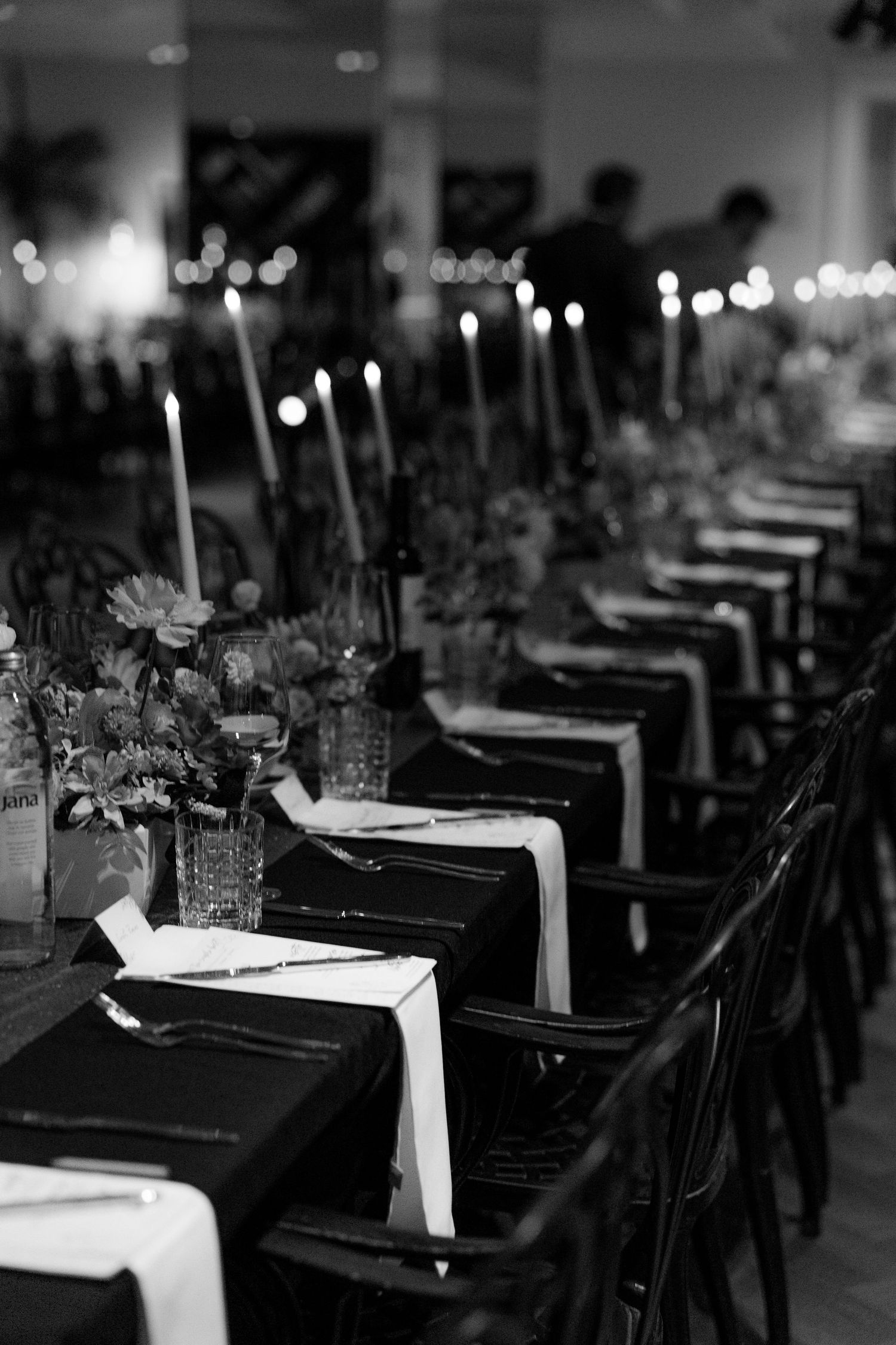 Black and white photo of elegant wedding reception tables set with candles, flowers, and dark napkins.
