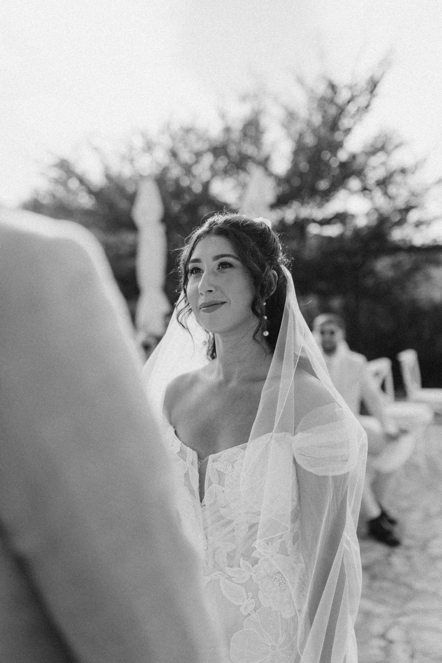 A bride wearing a white lace dress and veil sitting outdoors during her wedding ceremony.