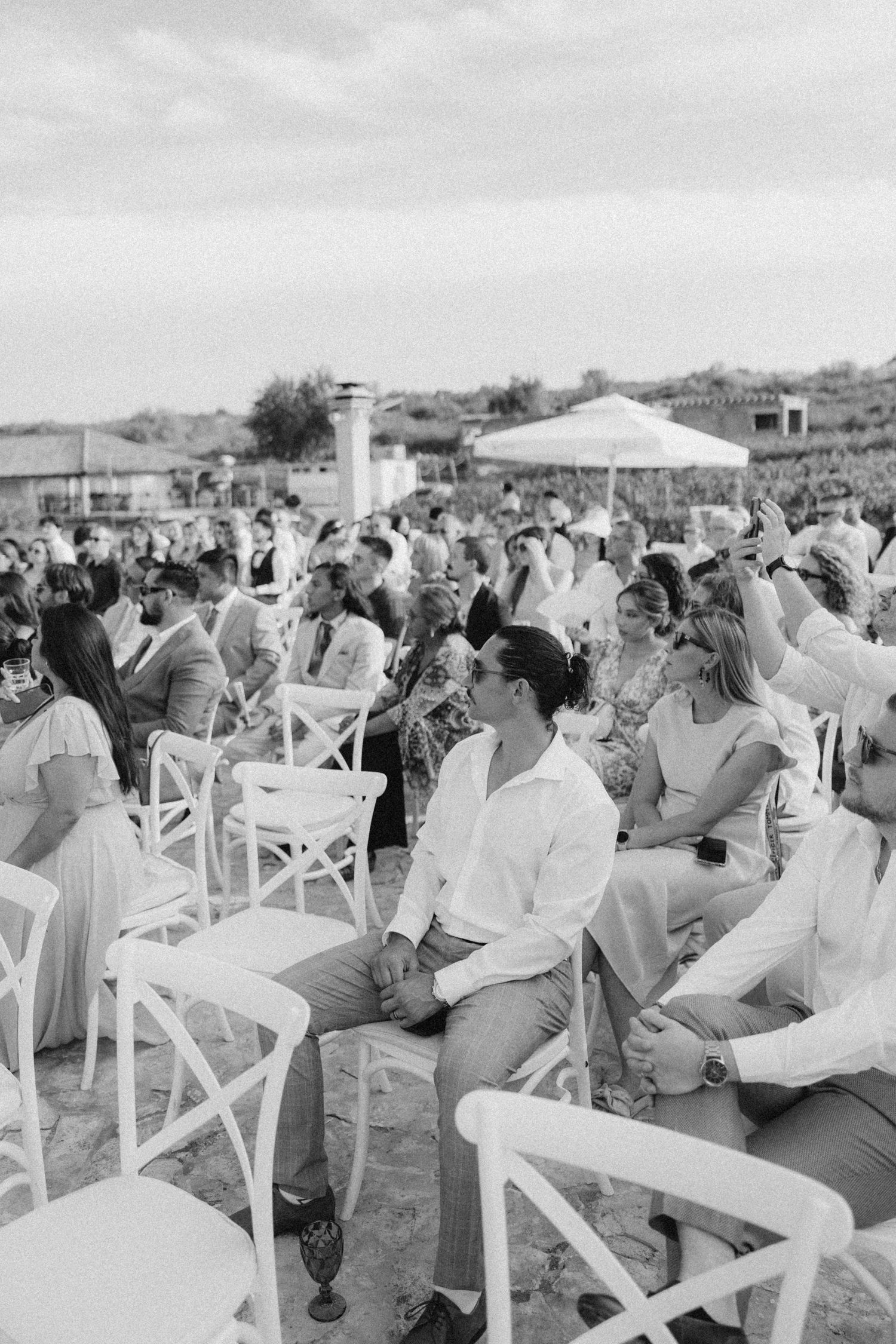 Black and white photo of wedding guests seated in white chairs at outdoor venue in Italy.