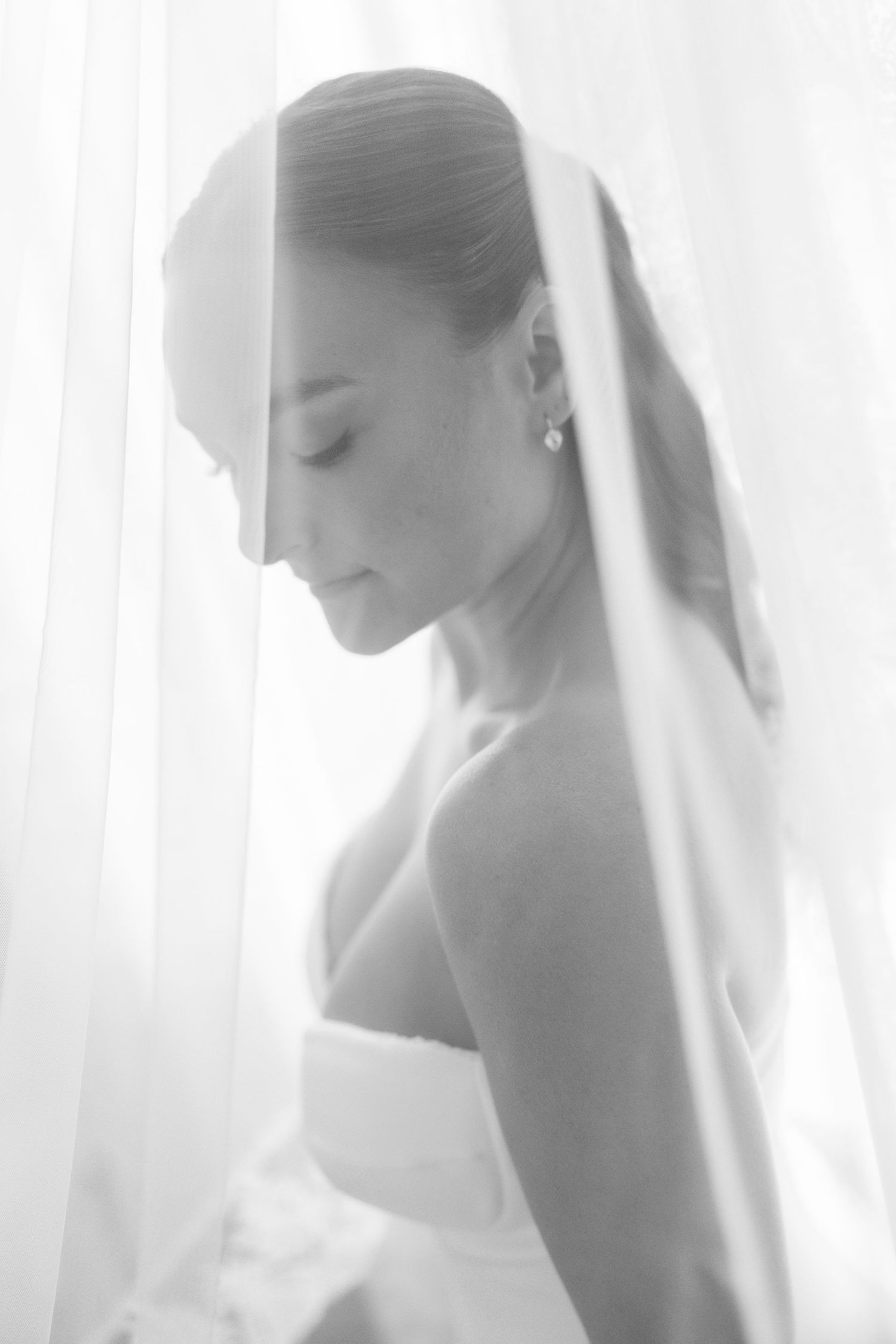 Dreamy black and white portrait of bride wearing veil looking down near sheer curtains.