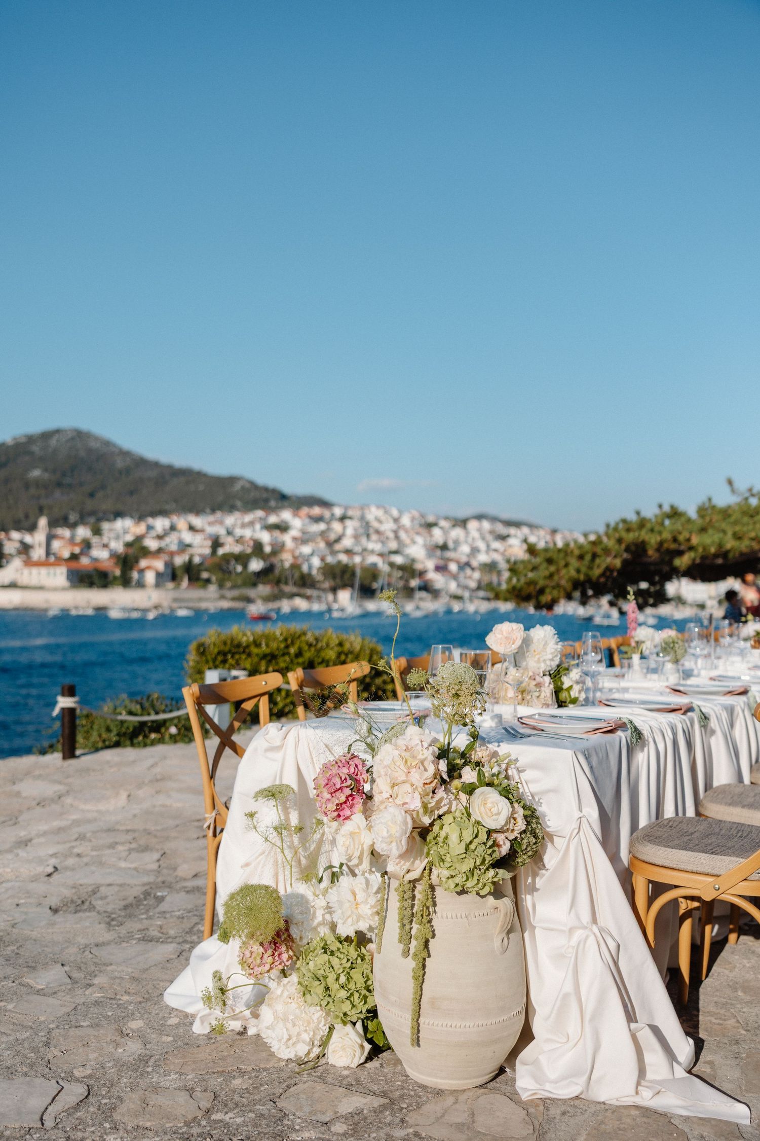 Outdoor waterfront wedding reception table with white floral arrangements and scenic coastal view.